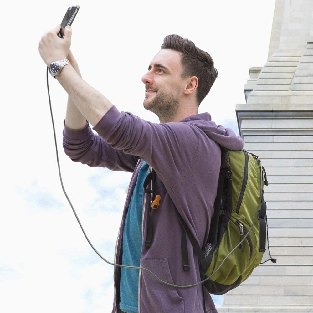 A man wearing a purple hoodie and green backpack smiles while taking a selfie with his phone connected to an Anker Powerline+ USB C to USB 3.0 Cable by Anker. He stands outdoors by a light stone building under a cloudy sky.