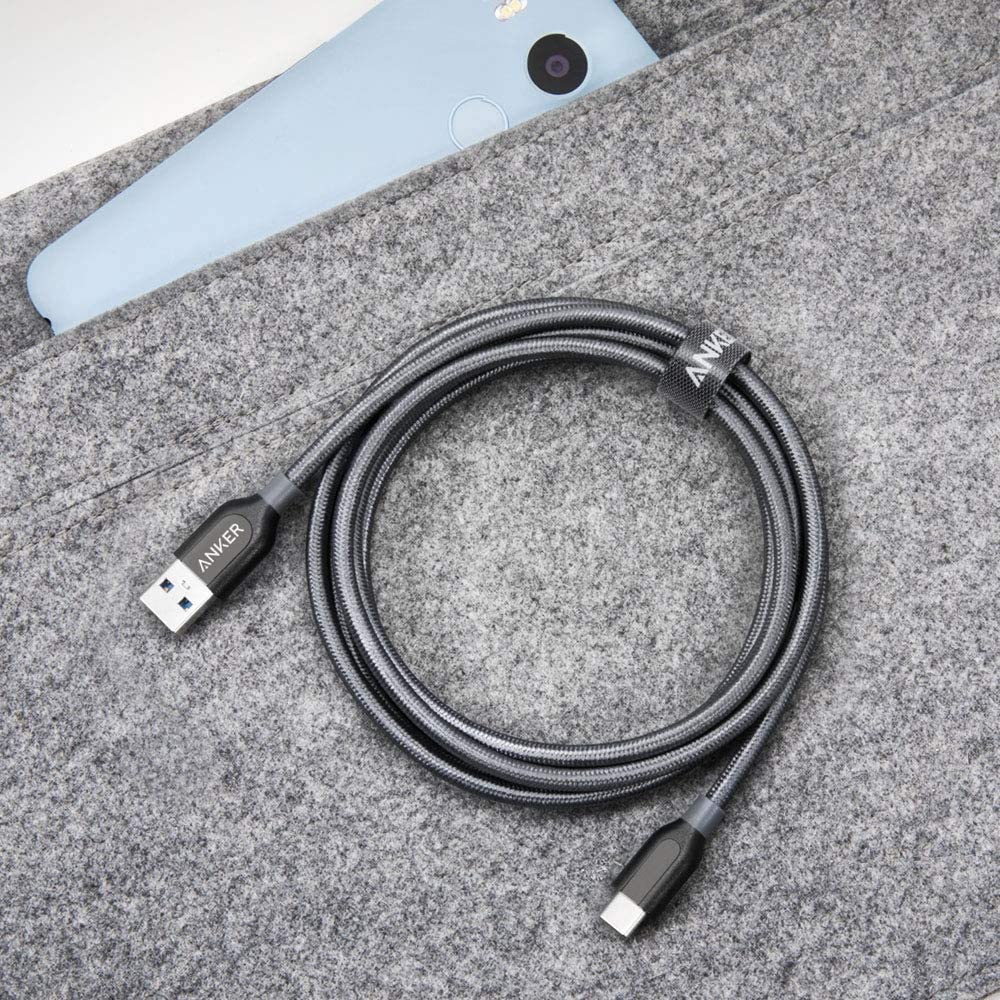 A gray braided Anker Powerline+ USB C to USB 3.0 Cable (3 ft, 6 ft) is coiled on a soft gray felt surface with “ANKER” branding. A light blue smartphone is partly visible from a matching pocket above the cable.