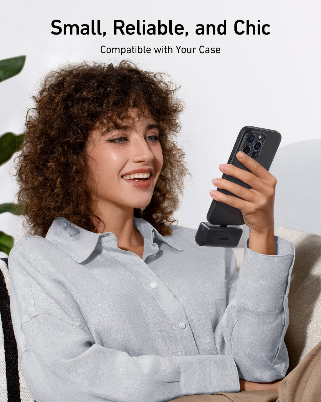 A smiling woman in a light gray shirt sits on a couch, using her smartphone powered by the Anker Nano Power Bank (22.5W, Built-In USB-C Connector) on a sleek black stand. Text above: "Small, Reliable, and Chic. Compatible with Your Case.