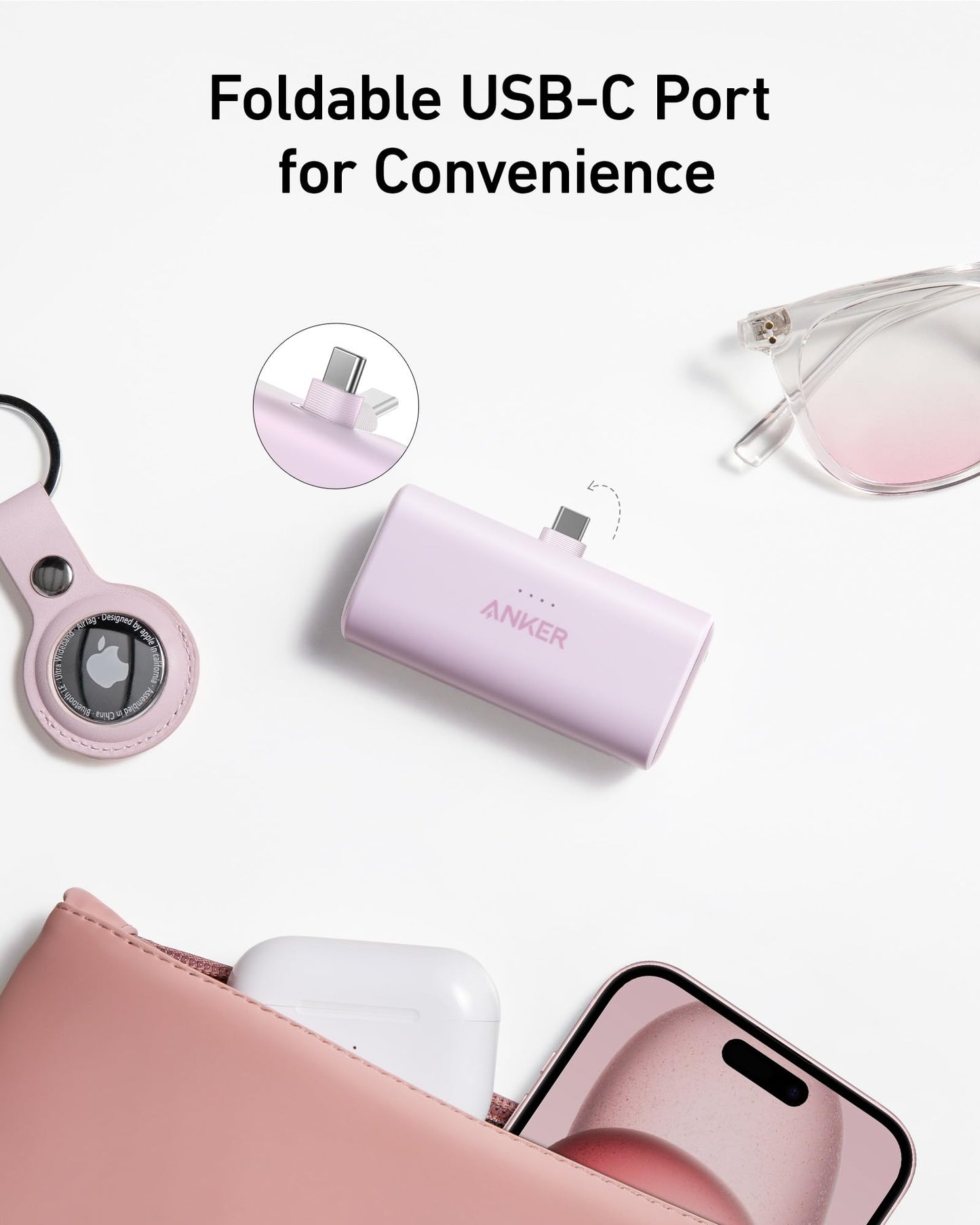 A pink Anker Nano Power Bank (22.5W, Built-In USB-C Connector) sits beside a pink pouch, AirPods case, iPhone, glasses, and an AirTag keychain. Text above says: Foldable USB-C Port for Convenience.