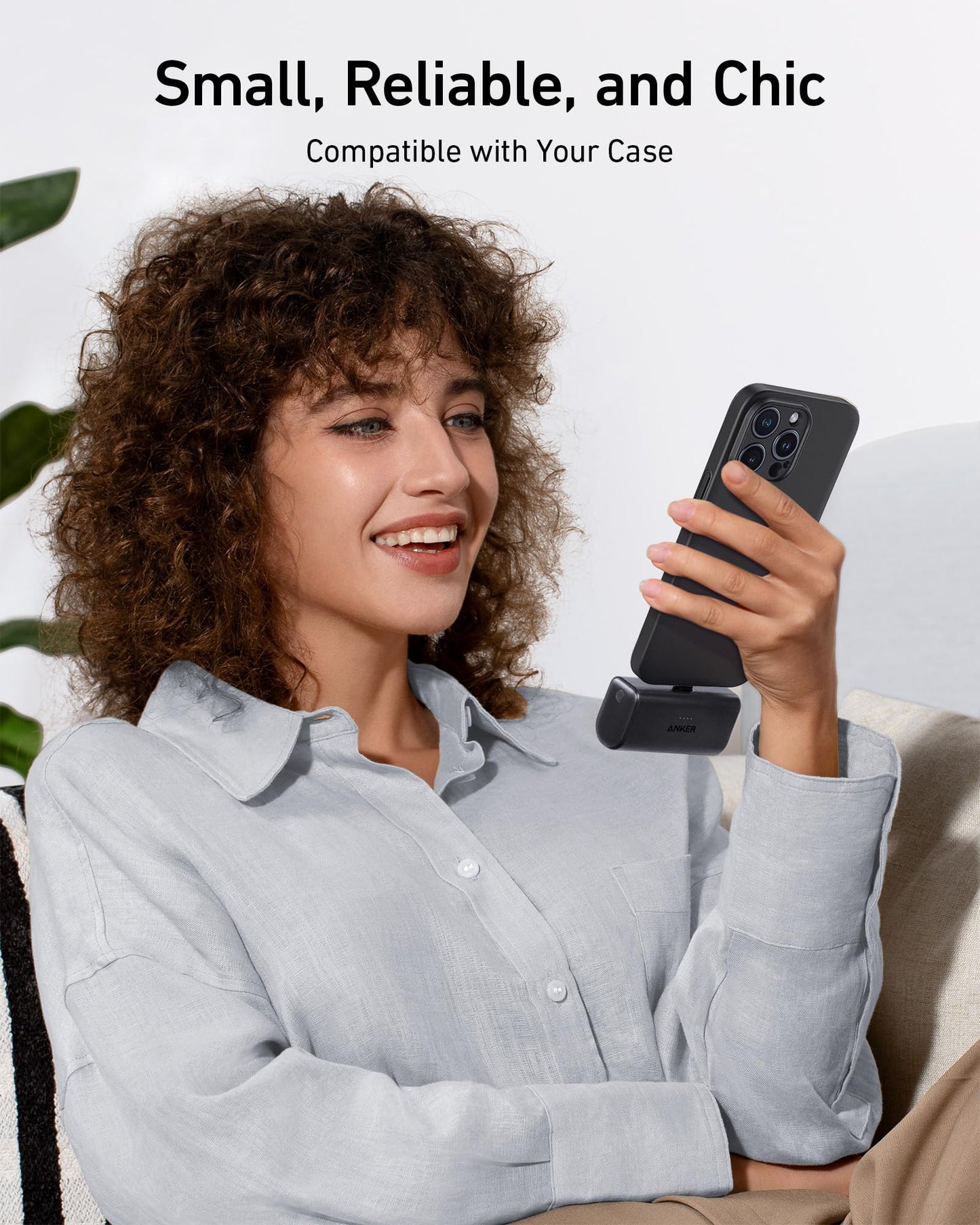 A smiling woman with curly brown hair, in a light gray button-up shirt, sits on a sofa holding her smartphone connected to the Anker Nano Power Bank (22.5W, Built-In USB-C Connector) by Anker. White walls and a plant are behind her. Text: Small, Reliable, and Chic.