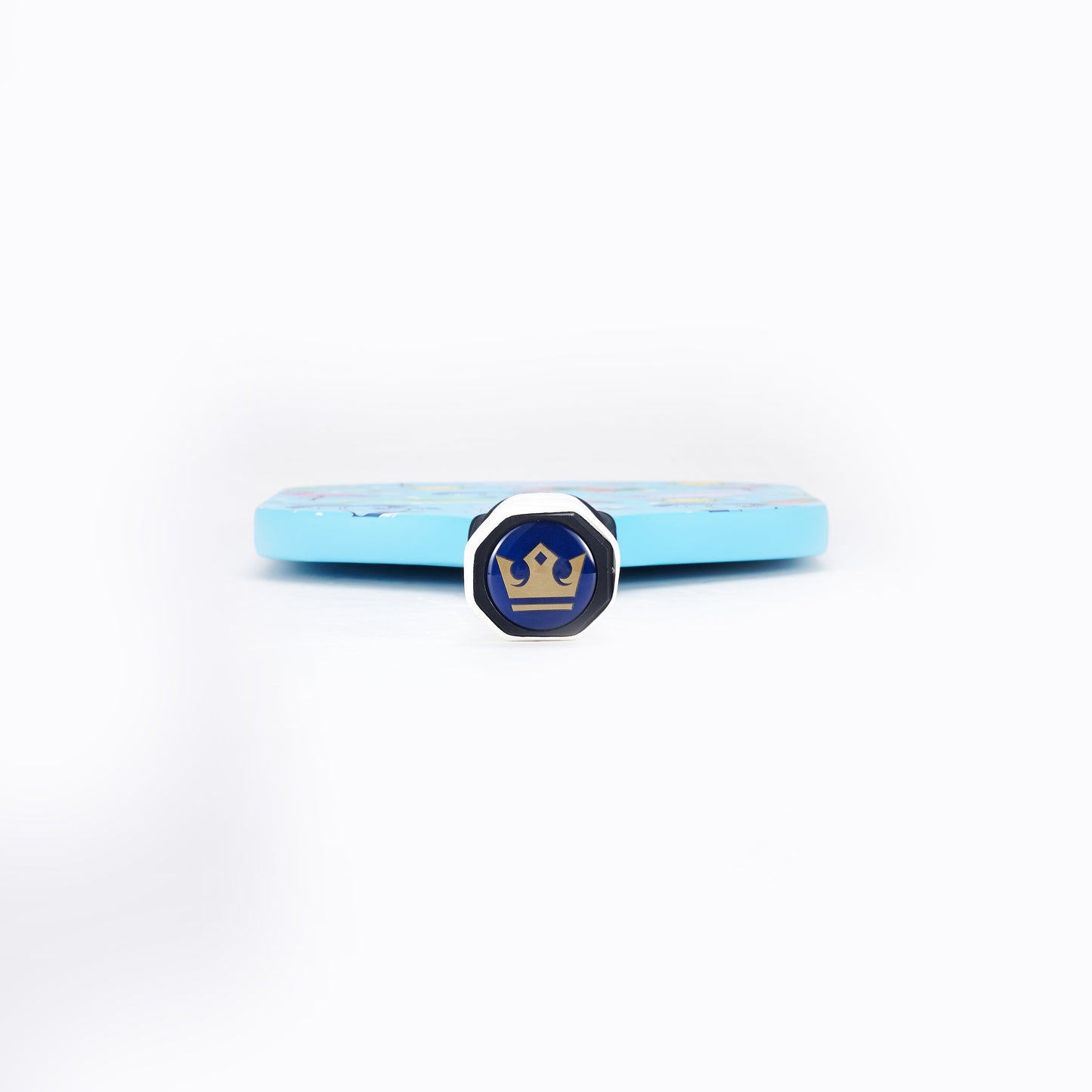 A silver hexagonal ring with a blue face and gold crown design stands upright before an ARTI Carbon Fiber "FLORIDA" Paddle by ARTI, which lies flat and blurred on a white background. The focus is on the ring.