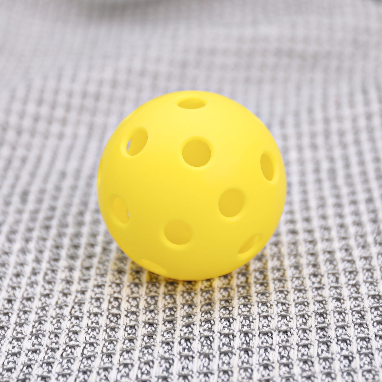 The ARTI Indoor Pickleball by ARTI is a bright yellow ball with evenly spaced round holes and a smooth finish, designed for durability and ideal for indoor play. It sits on a textured gray-and-white patterned fabric surface.