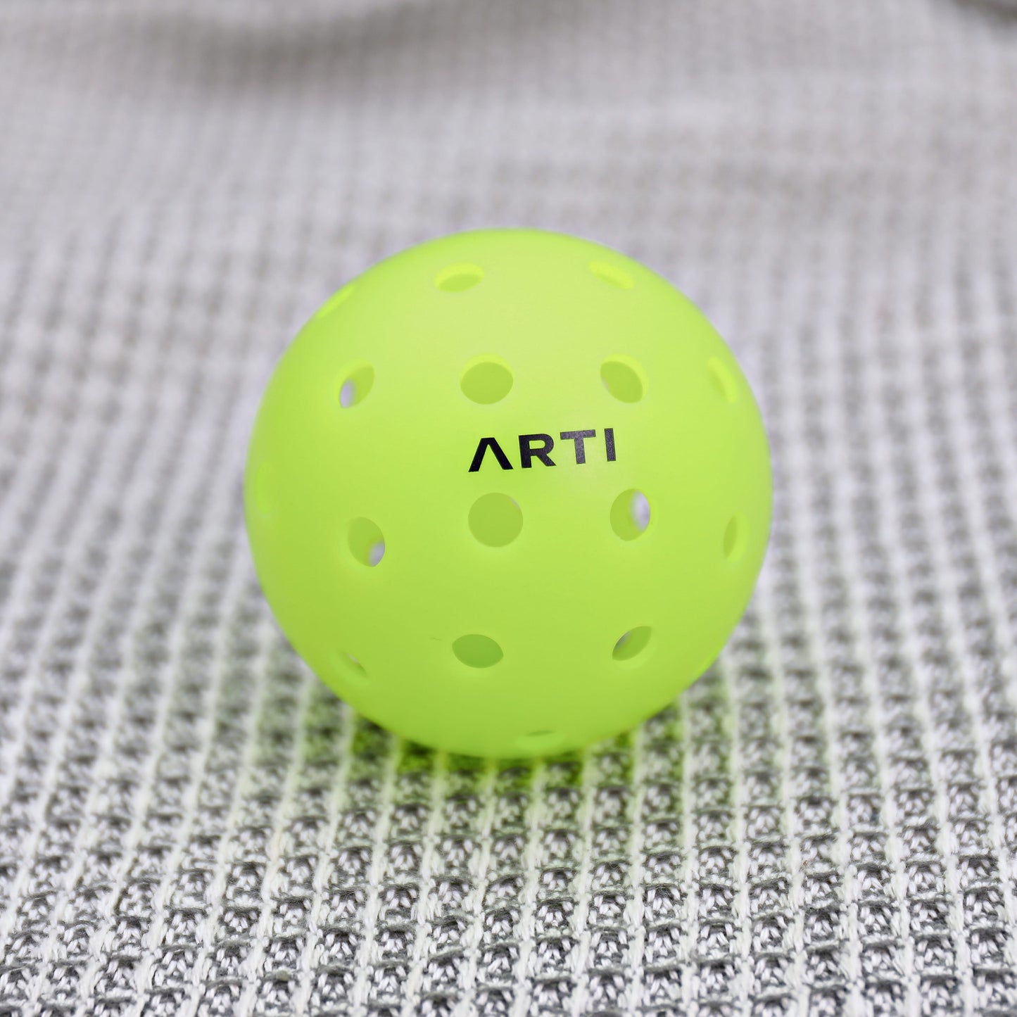 A bright yellow-green ARTI Outdoor Pickleball, featuring the ARTI brand name and evenly spaced holes for durability, rests on a gray and white woven fabric surface.