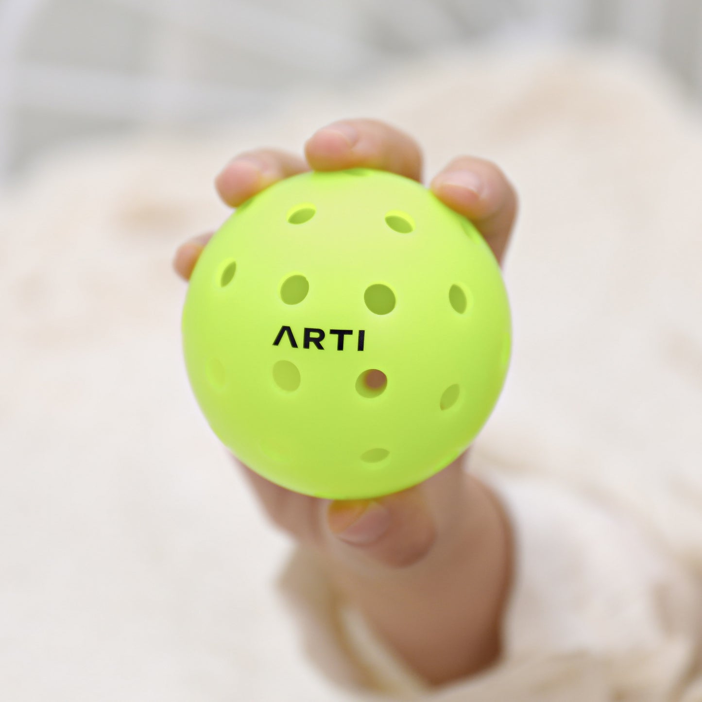 A small hand holds the ARTI Outdoor Pickleball, a bright yellow ball with evenly spaced holes and "ARTI" printed in bold black letters, standing out against a soft, blurred background.