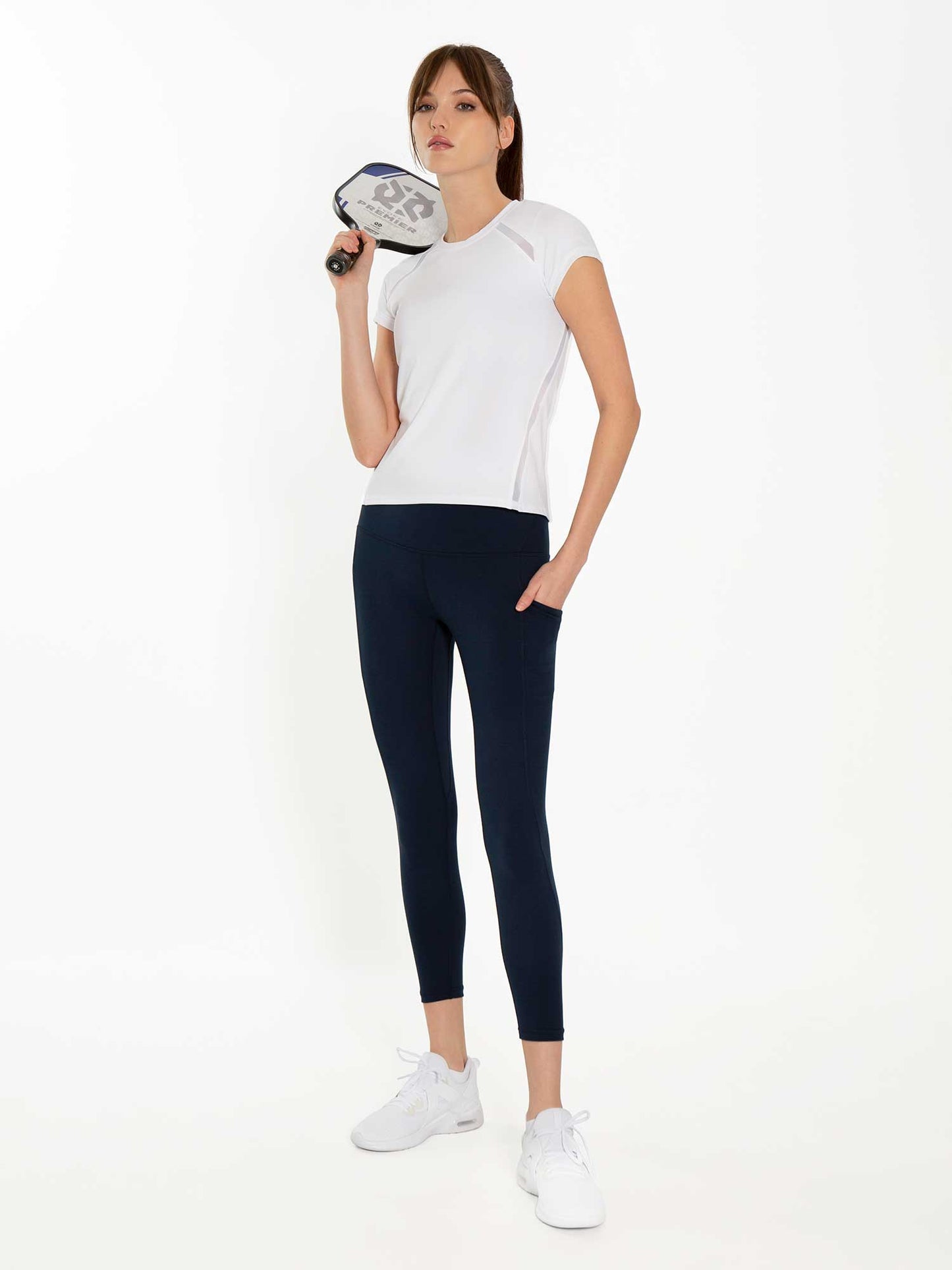 A woman confidently poses against a white background, holding a pickleball paddle. She wears the inPhorm NYC Ashley 7/8 Legging in Midnight, paired with a white short-sleeve athletic top and white sneakers. Her hair is tied back.