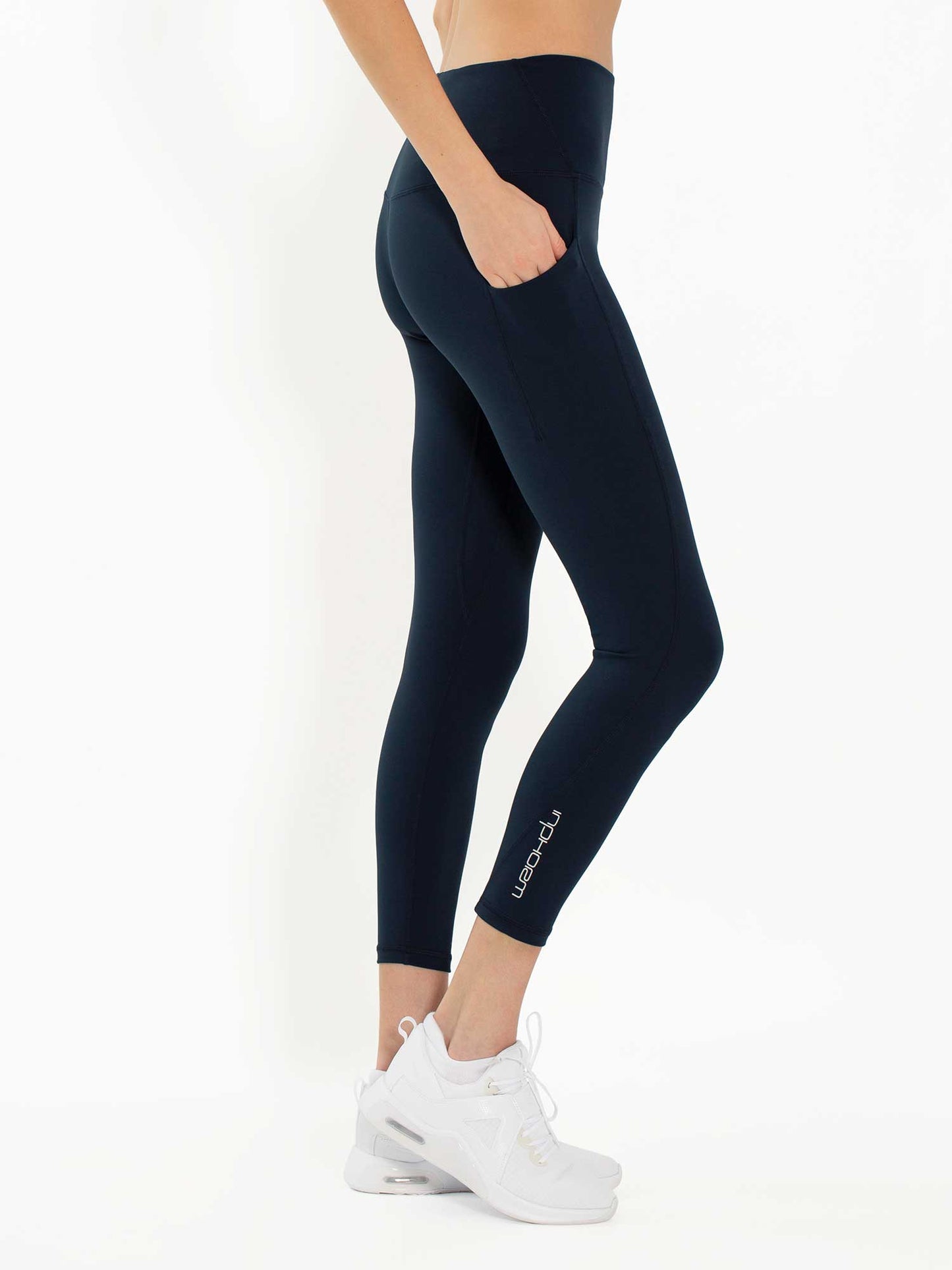 A person, visible from the waist down, wears inPhorm NYC Ashley 7/8 Legging - Midnight: high-waisted, dark navy leggings with four-way stretch, a side pocket, and white sneakers. The brand name appears near the left ankle. The background is white.