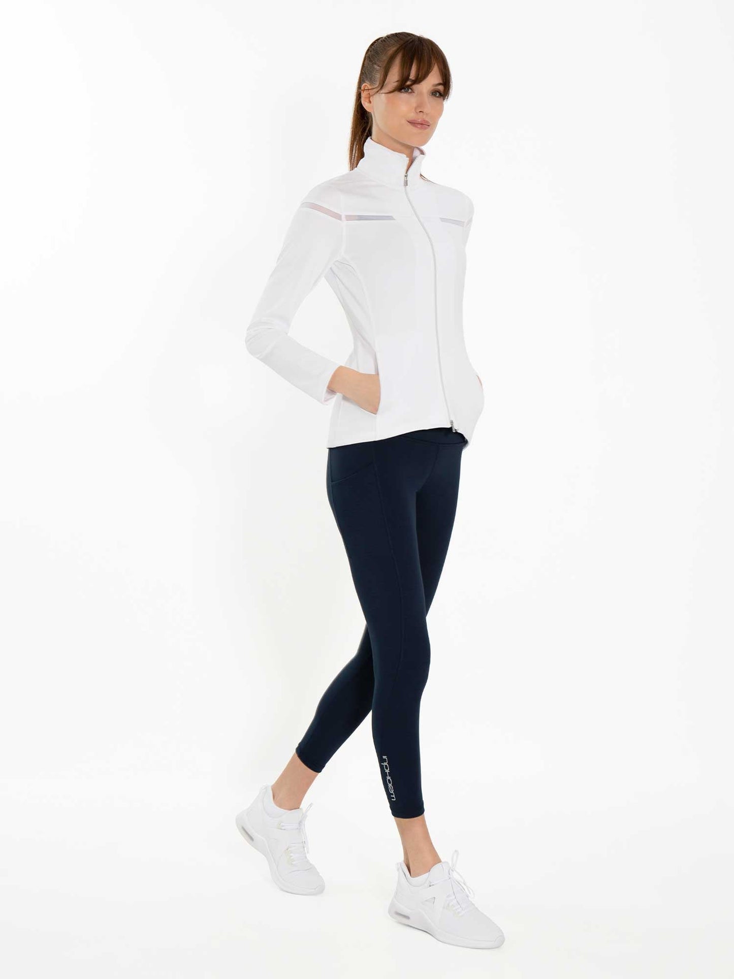 A woman stands against a white background, wearing inPhorm NYC’s Ashley 7/8 Legging in Midnight, a fitted white zip-up jacket, and white athletic shoes. Her hands are in her jacket pockets, and her brown hair is pulled back in a ponytail.