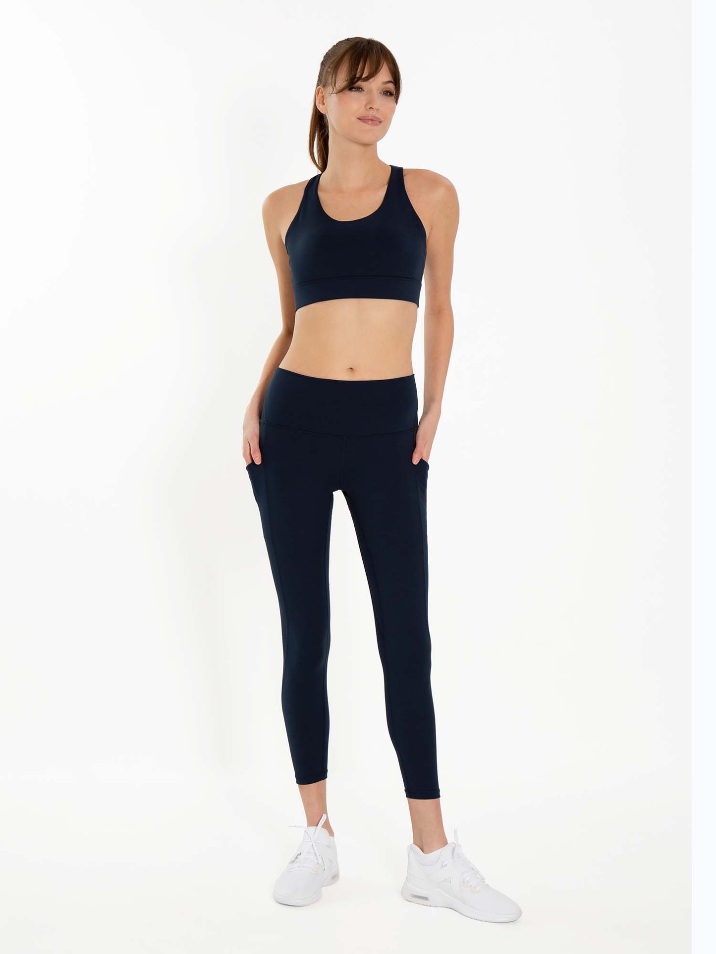 A woman with light skin and straight brown hair in a ponytail wears the inPhorm NYC Ashley 7/8 Legging - Midnight and a matching navy sports bra. She stands against a white background, hands in her pockets, looking slightly to the side.