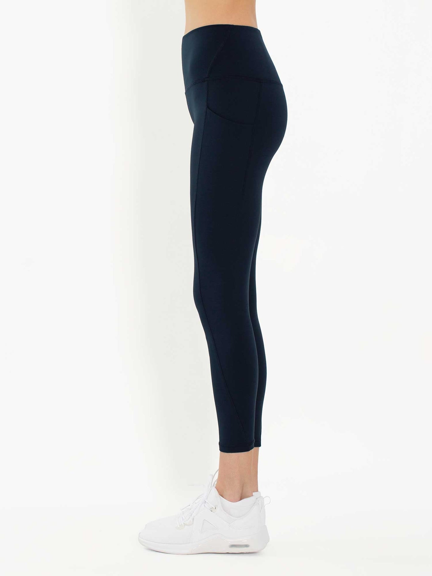 A person stands in profile against a white background, showing only the lower torso and legs, wearing inPhorm NYC's Ashley 7/8 Legging - Midnight with four-way stretch comfort, ending just above the ankles, paired with white sneakers.