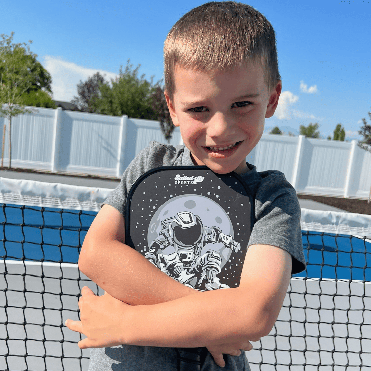 A young boy smiles while hugging his Salted City Sports Astro | Junior Series Kids Pickleball Paddle, featuring an astronaut and moon design. He stands on an outdoor court with a net, blue surface, and bright sky in the background.