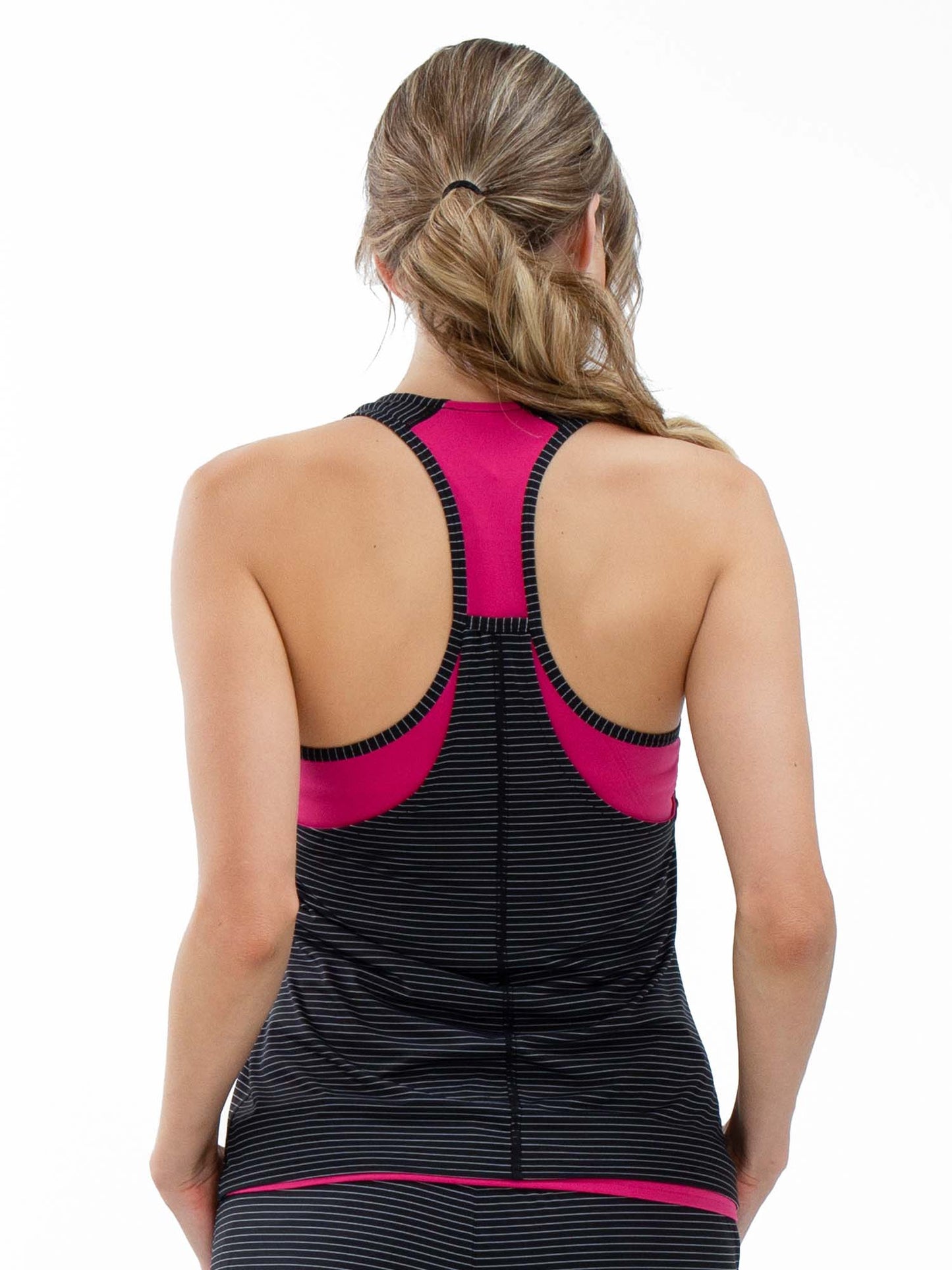 A woman with light brown hair in a low ponytail is seen from the back wearing the inPhorm NYC Aubrey Double Layer Athleisure Tank in Fuchsia/Black, featuring thin stripes, vibrant pink panels, black trim, and soft nylon fabric.