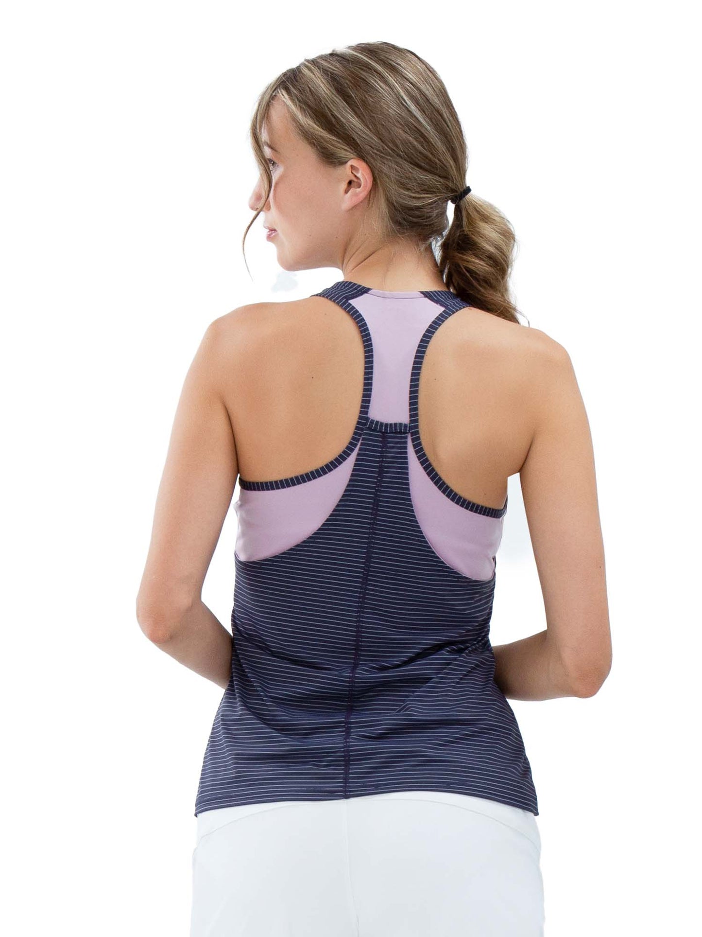 A woman with light brown hair in a low ponytail is seen from the back, wearing the inPhorm NYC Aubrey Double Layer Athleisure Tank in Lilac/Dark Shadow with white bottoms, against a plain white background, looking to her left.
