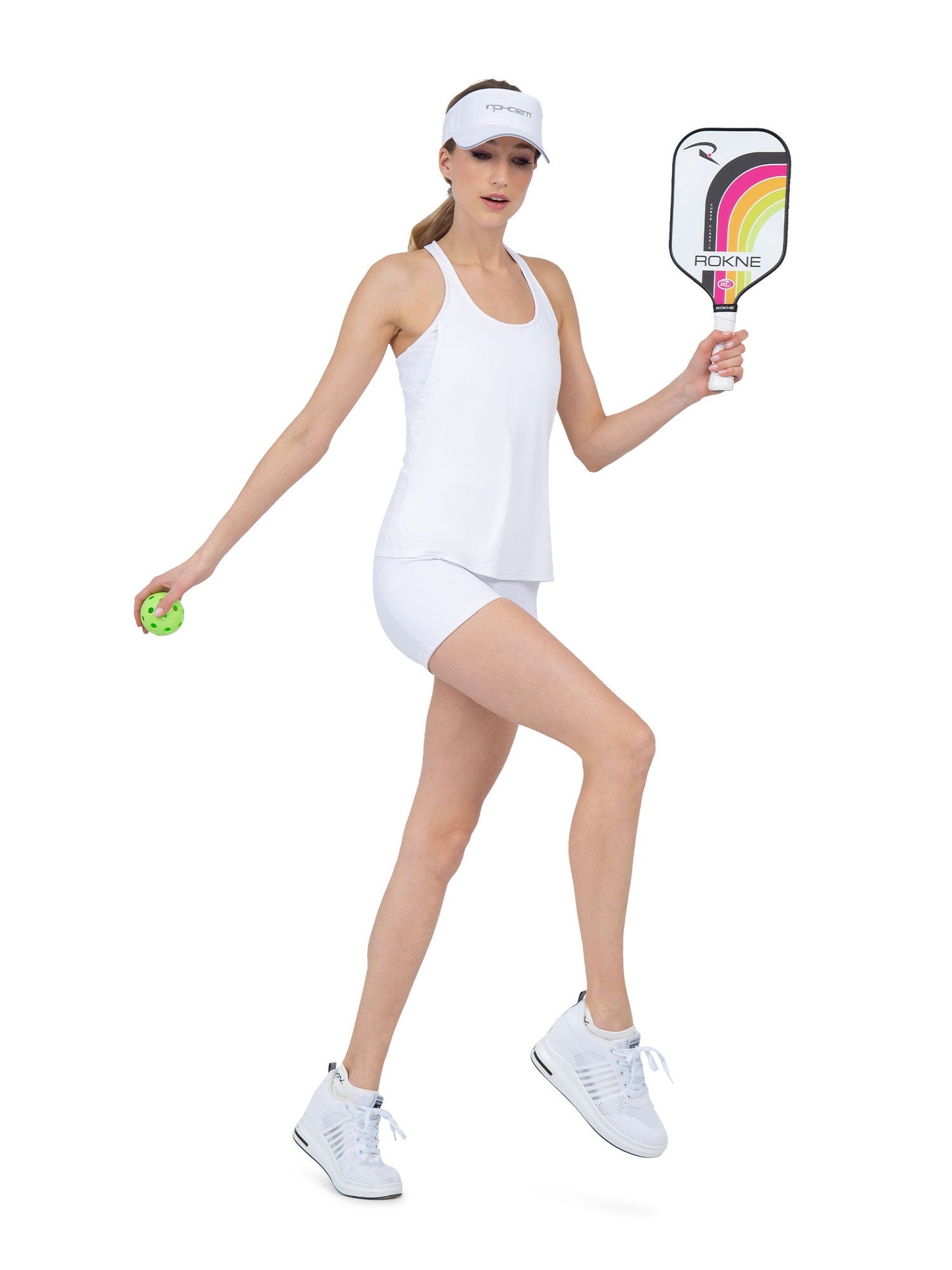 A woman in all-white athletic wear, featuring the inPhorm NYC Aubrey Double Layer Athleisure Tank in White, stands on a white background holding a green pickleball and colorful paddle, ready to serve. She also wears sneakers, shorts, and a visor.