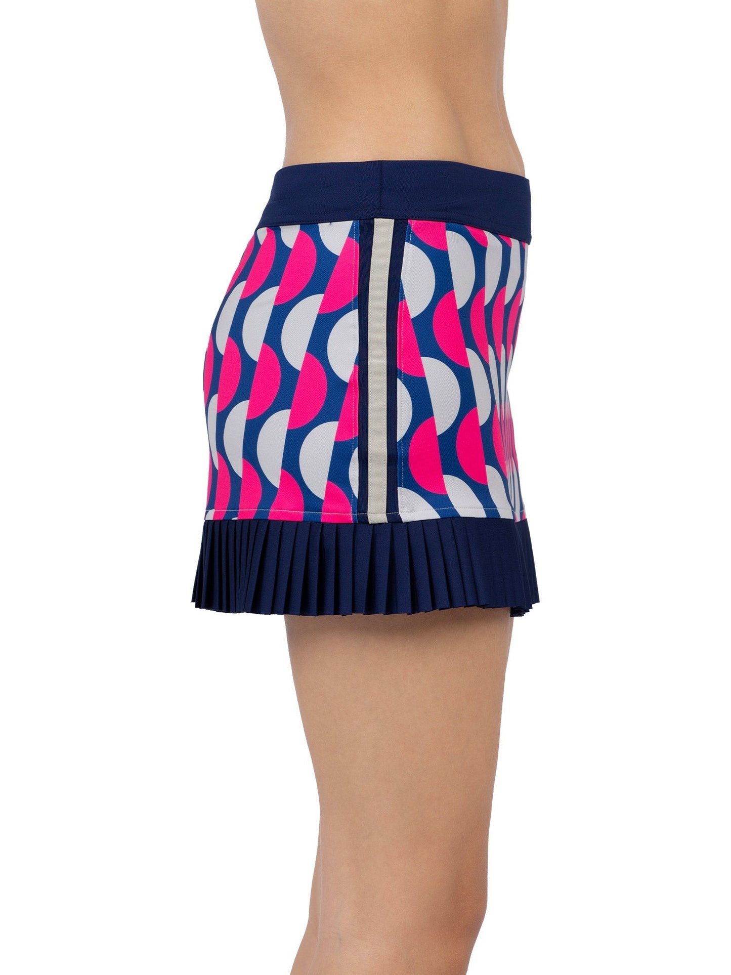 A person models the Audrey Tennis & Pickleball Skirt by inPhorm NYC, featuring bold pink, blue, and white geometric pleats, a dark blue waistband, vertical beige side stripe, and dark blue hem. Only the midsection and thighs are visible.