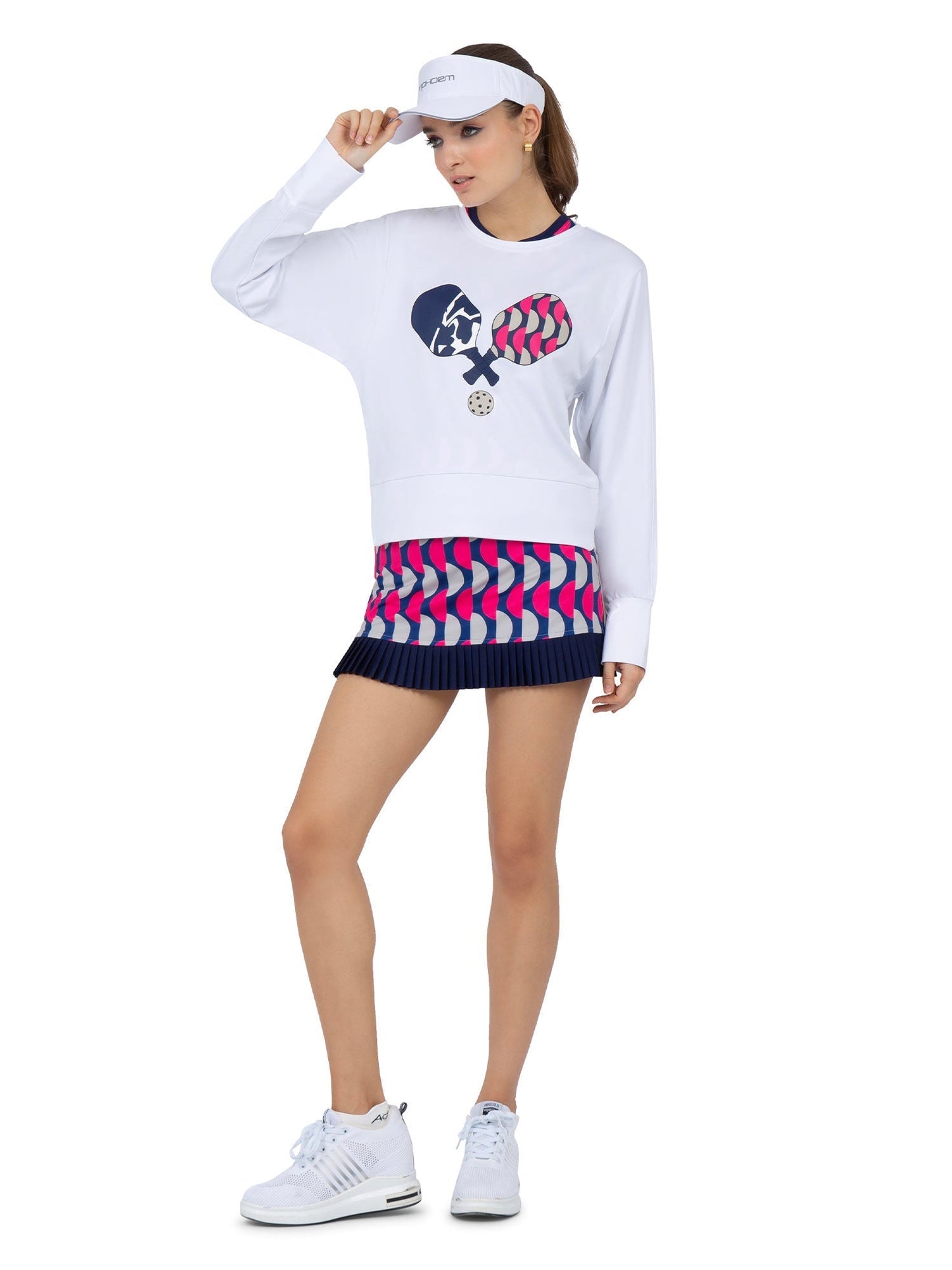 A woman models the inPhorm NYC Audrey Tennis & Pickleball Skirt—navy, pink, and white geometric pleats—paired with a white visor, sneakers, and a tennis graphic sweatshirt against a white backdrop.
