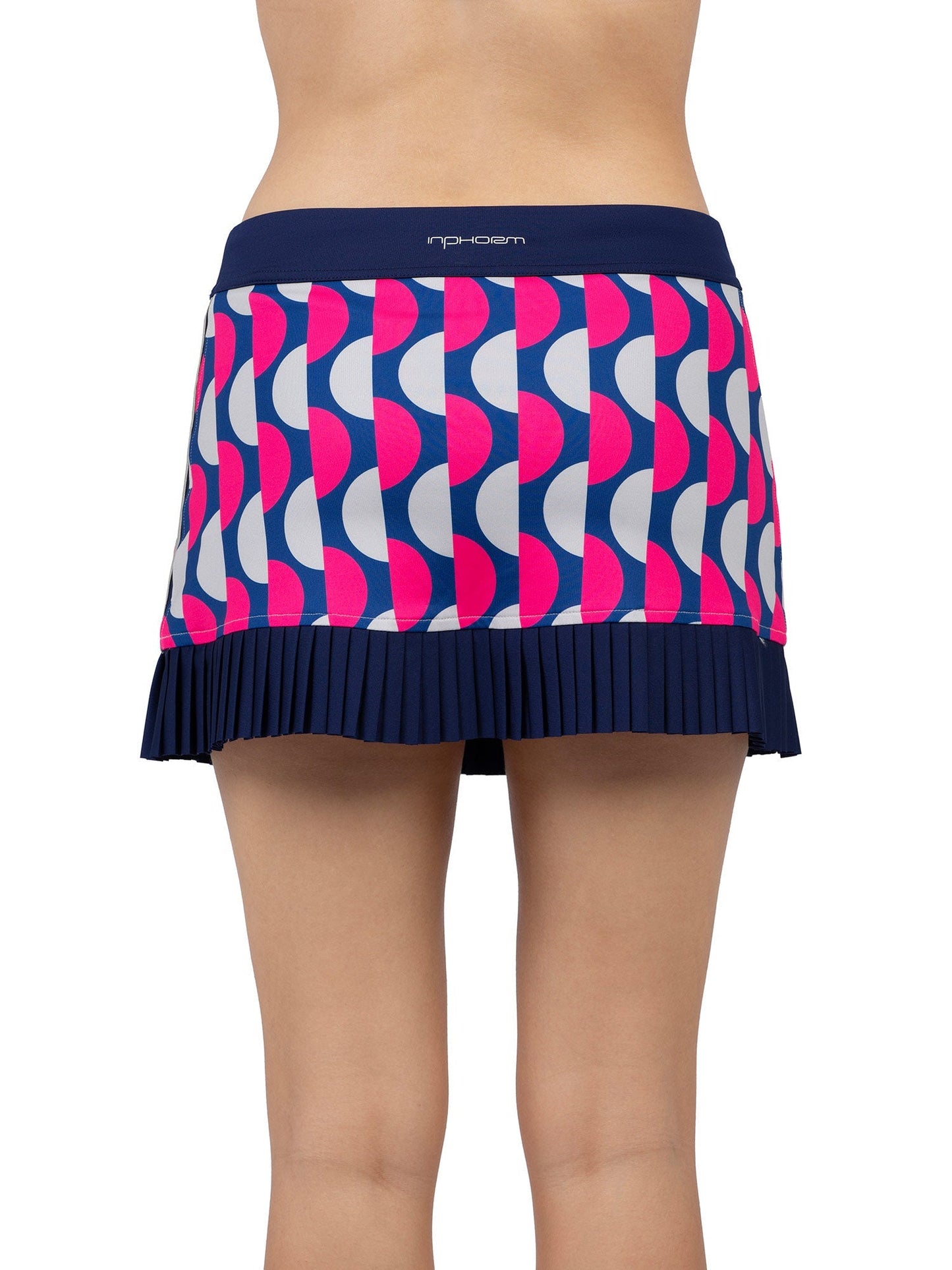 A person stands facing away, showing only the lower back and legs, wearing the inPhorm NYC Audrey Tennis & Pickleball Skirt—a navy pleated skirt with bold red and white geometric circles, a solid waistband, and a finely pleated hem for a stylish look.