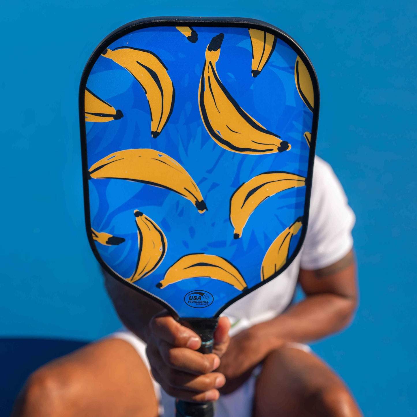 Dressed in white, a person sits before a bright blue background, holding up the Swinton Pickleball Hero Bananas Pickleball Paddle, its bold banana design and USA Pickleball approved graphite face playfully concealing their face.