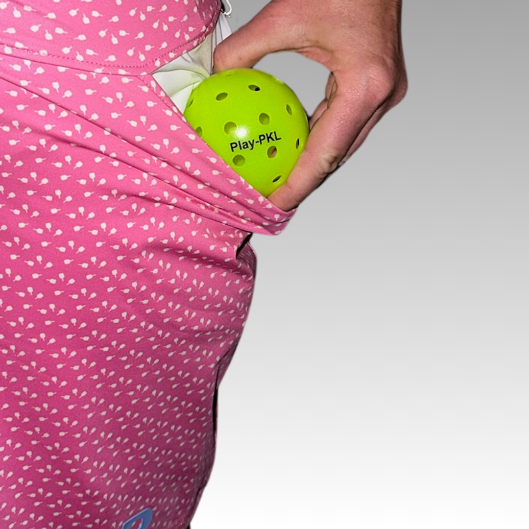A hand places a bright green Banger Hard Surface Pickleball by Playly Store into the pocket of a pink skirt with white hearts, set against a simple gray gradient background. Perfect for indoor play.
