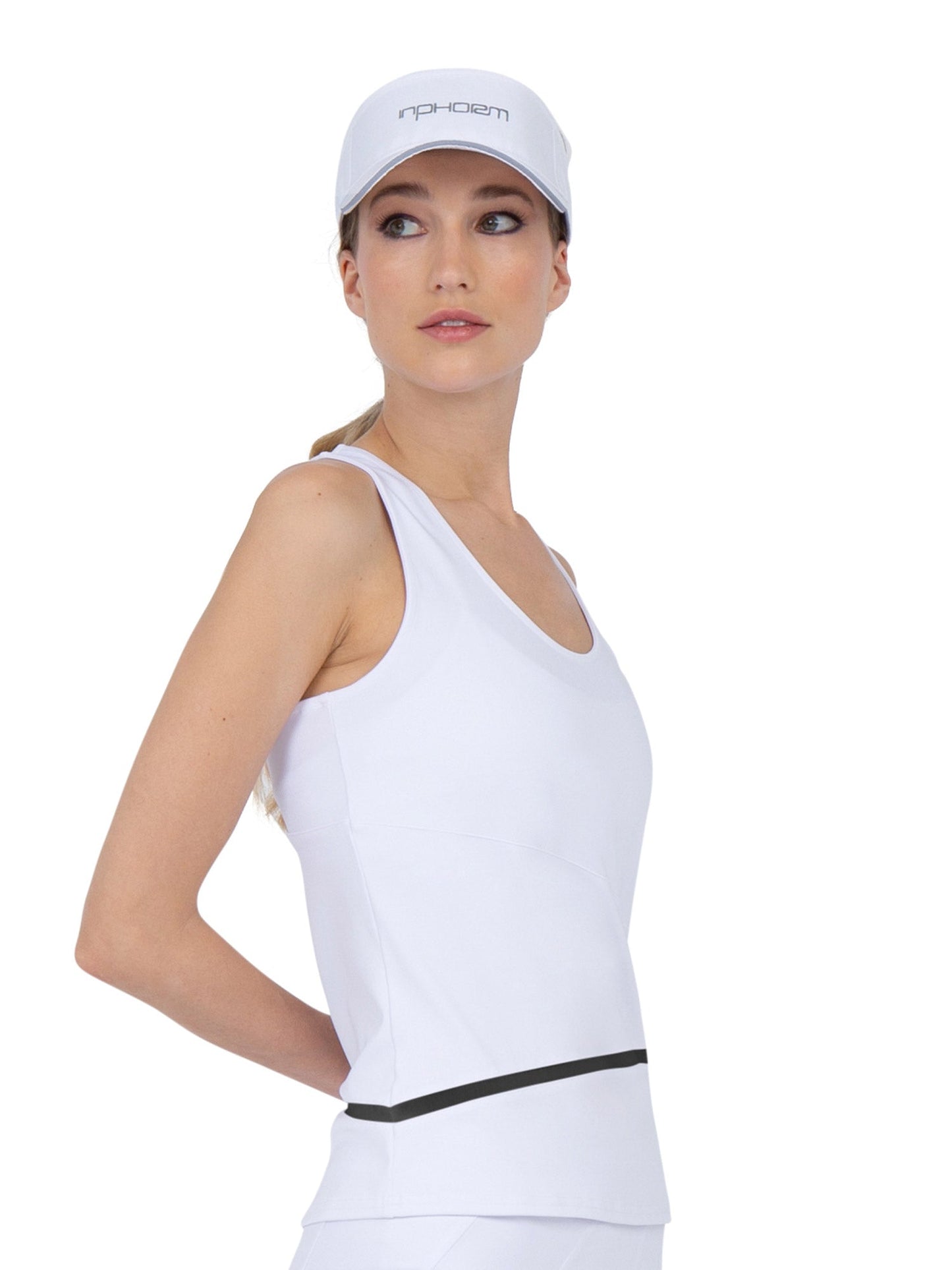 A woman wears the inPhorm NYC Baseline Tank in White/Black and a matching skirt, plus a white baseball cap with front text, standing sideways and looking over her shoulder against a neutral background.