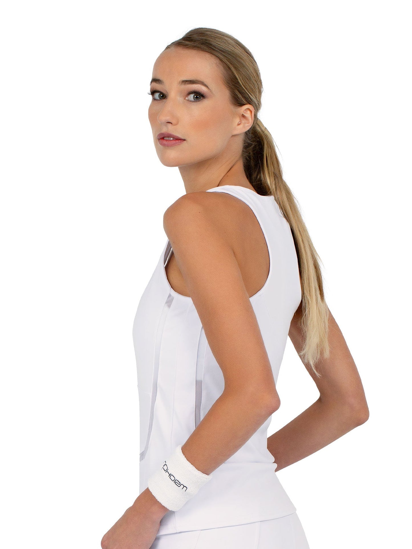 A woman with long blonde hair in a low ponytail models the inPhorm NYC Baseline Tank - White and a matching wristband, standing against a plain white background and looking over her shoulder with a neutral expression.