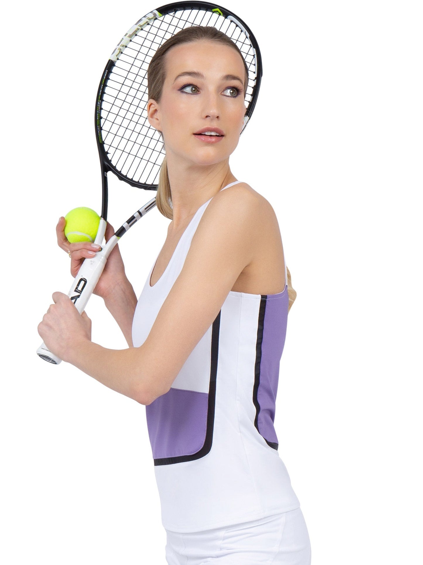 A woman in the inPhorm NYC Baseline Tank - White/Lavender Combo holds a racket and yellow tennis ball, ready to serve. She has light skin, blonde hair tied back, and looks to the side against a plain white background.