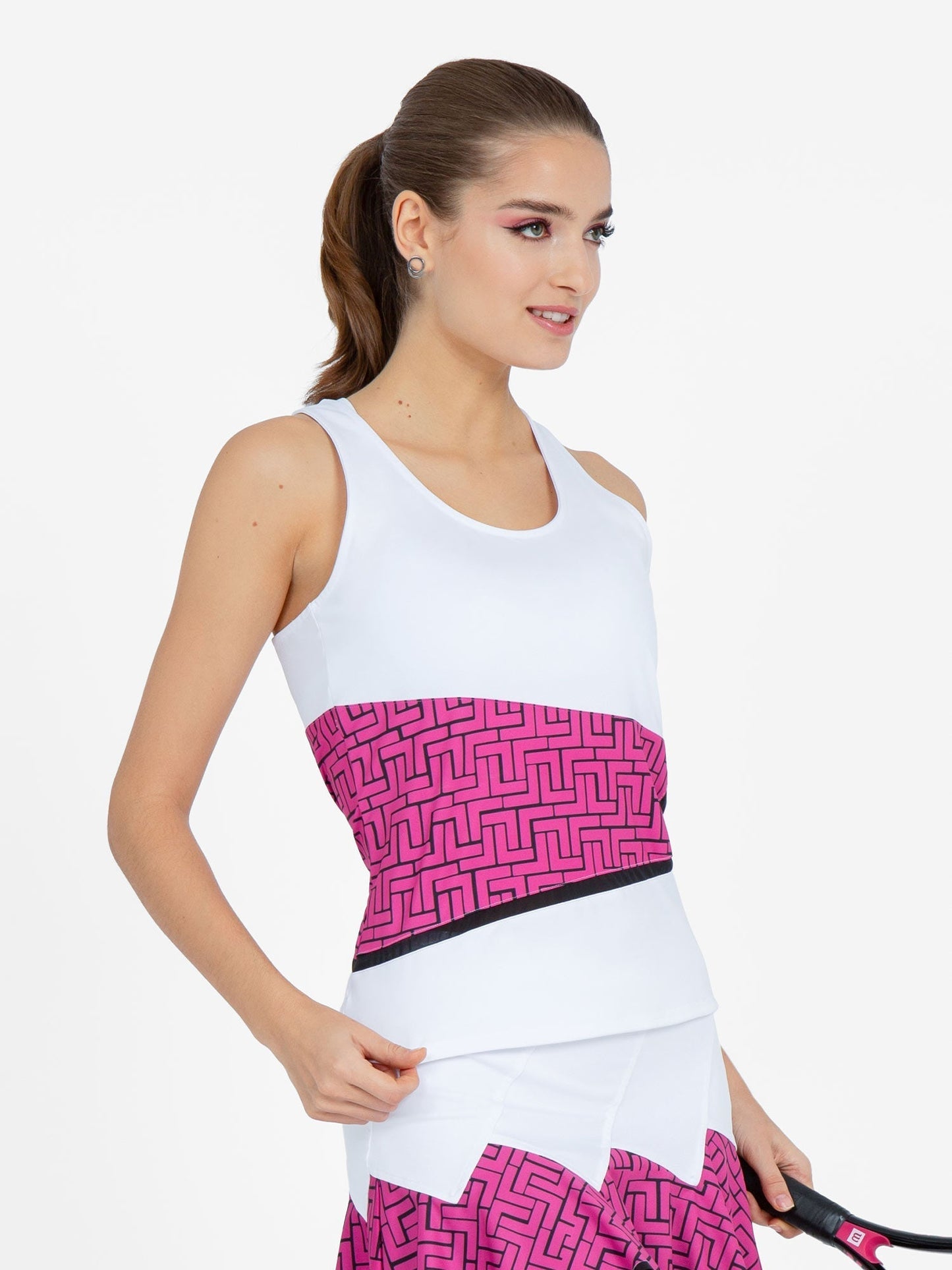 A woman with long brown hair in a ponytail wears the inPhorm NYC Baseline Tank in White/Pink Tessel, smiling slightly and holding a tennis racket against a plain white background.