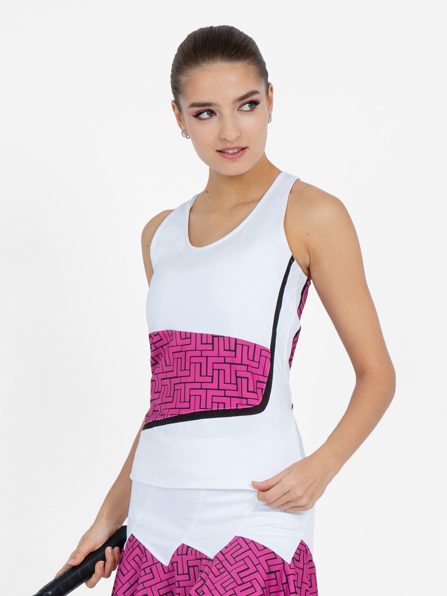 A woman wears the inPhorm NYC Baseline Tank in White/Pink Tessel, featuring a pink geometric waist pattern. With her hair in a neat bun, she holds a tennis racket by her side and looks right, set against a plain white background.