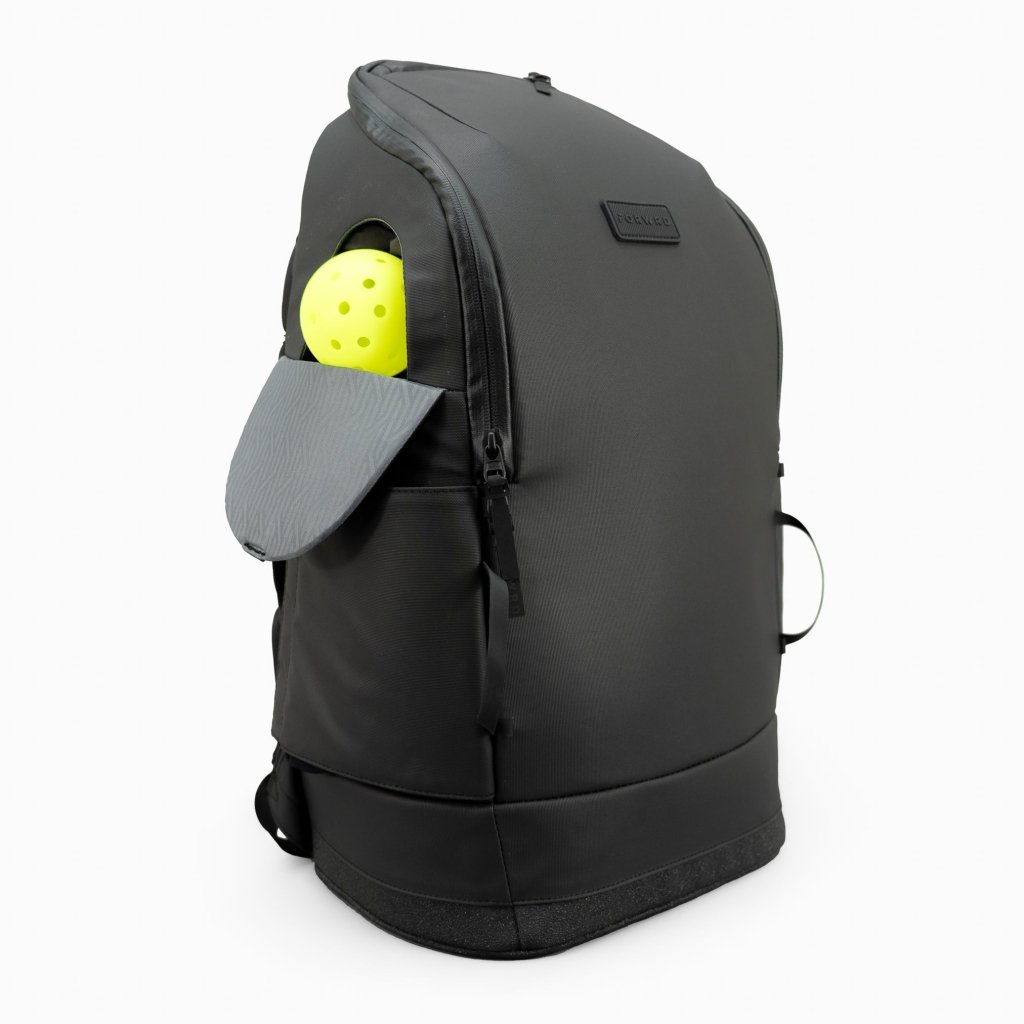 The FORWRD Court Caddy Pickleball Bag stands upright on a white background, with its side pocket open to reveal a gray cap and bright yellow pickleball. Made of premium materials, it has a small rectangular brand patch near the top.