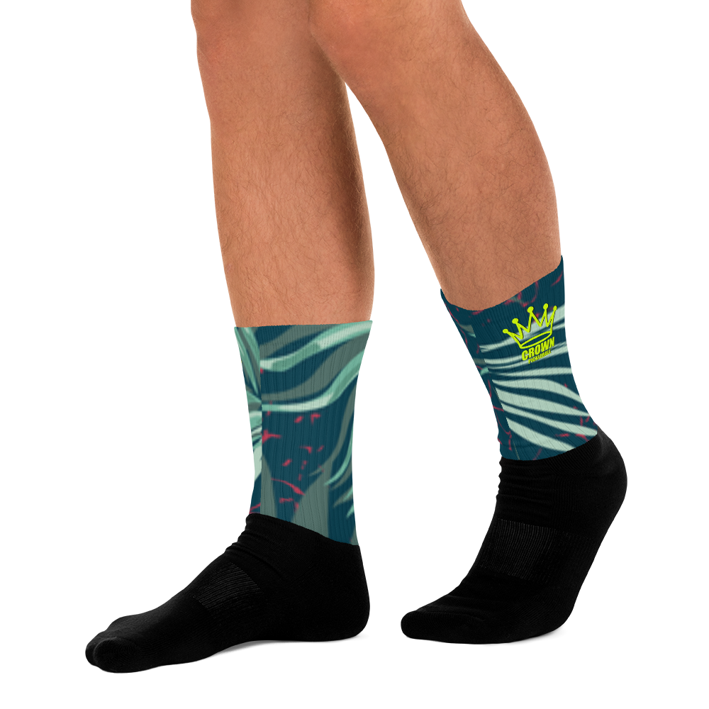 A person with light to medium skin wears Crown Pickleball's CROWN PICKLEBALL - Tropical Socks (Green), crew length socks featuring green tropical leaf patterns, pink accents, and a yellow crown "ORIGINAL" logo. The background is plain white.