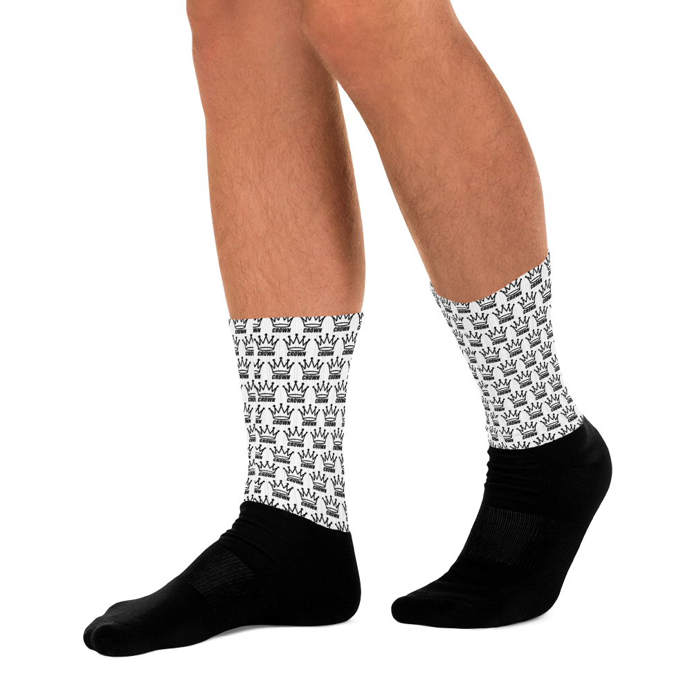 A person wears Crown Pickleball’s CROWN PICKLEBALL - Checkered Crown (White) crew socks, featuring black feet and white shafts with a repeating black crown pattern. Only their lower legs and feet are shown against a light gray background.