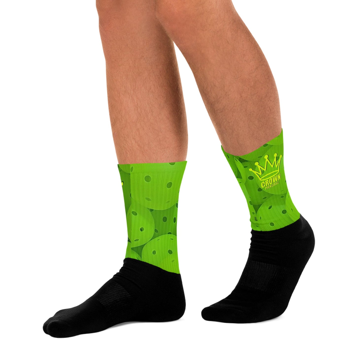 A person wears Crown Pickleball’s Performance Athletic Crew Socks, featuring black feet, bright green tops with round designs, and a yellow CROWN logo. Legs are shown from mid-thigh to floor on a white background.