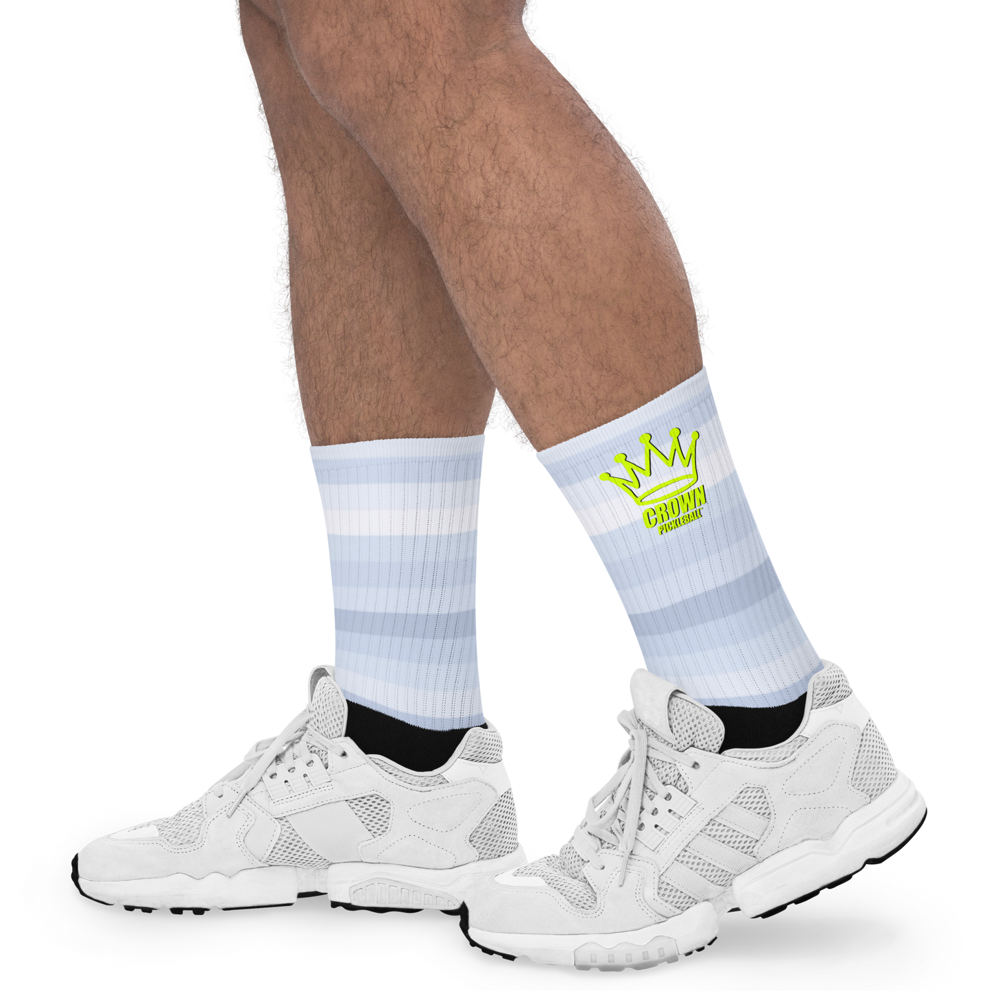 A person wears Crown Pickleball’s CROWN PICKLEBALL Performance Athletic Crew Socks—light blue and white striped socks with a yellow crown graphic and "CROWN" text—paired with white sneakers. Only their legs and feet are visible against a white background.