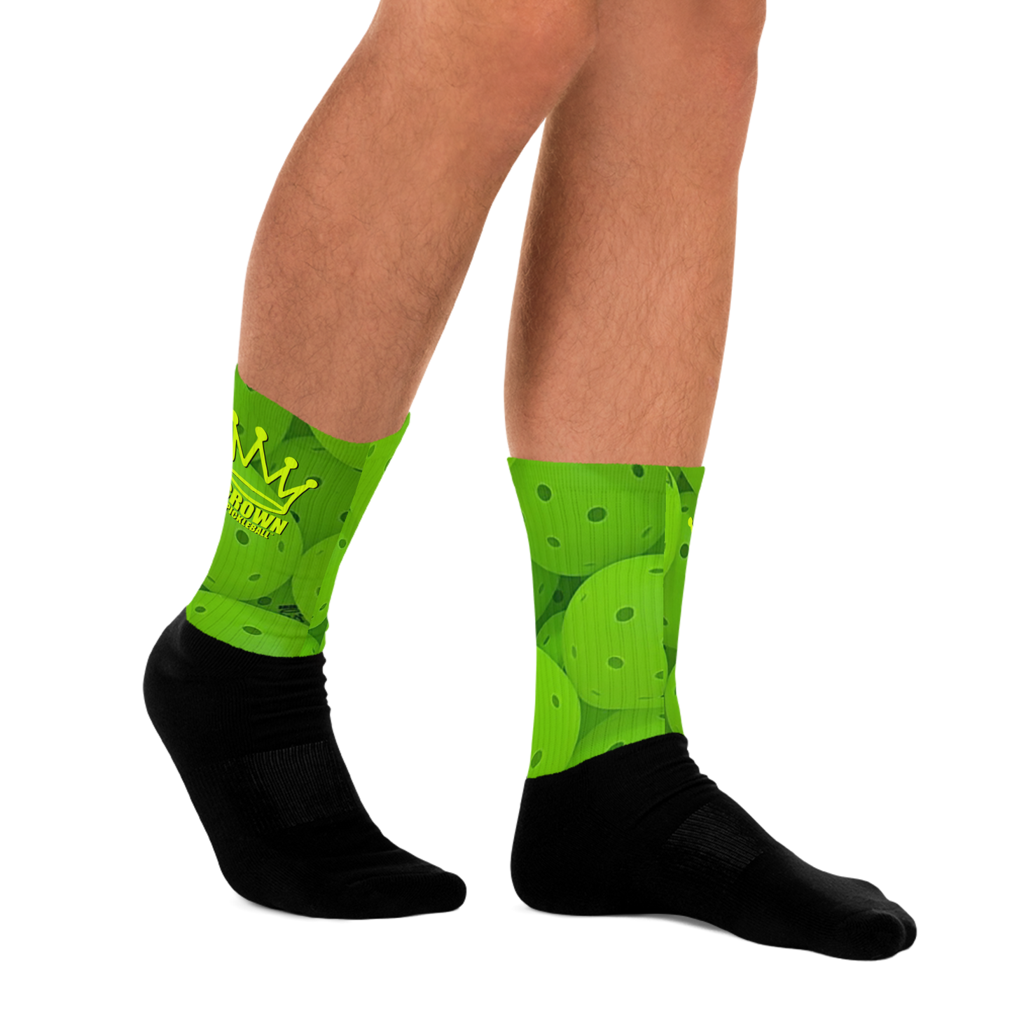 A person wears Crown Pickleball’s CROWN PICKLEBALL - Pickleball 2.0 Socks (Lime): crew-length, with lime pickleball paddles and balls on the calf, solid black foot and heel, plus a yellow crown and PICKLETOWN text on one sock.