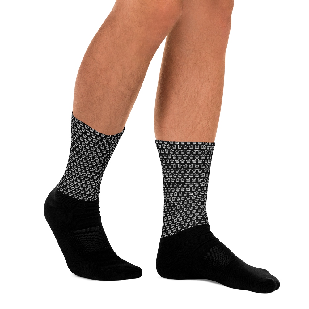 A person stands on a light gray surface wearing Crown Pickleball's CROWN PICKLEBALL - Checkered Crown (Black) crew socks with a geometric white pattern, visible from mid-calf down against a plain white background.