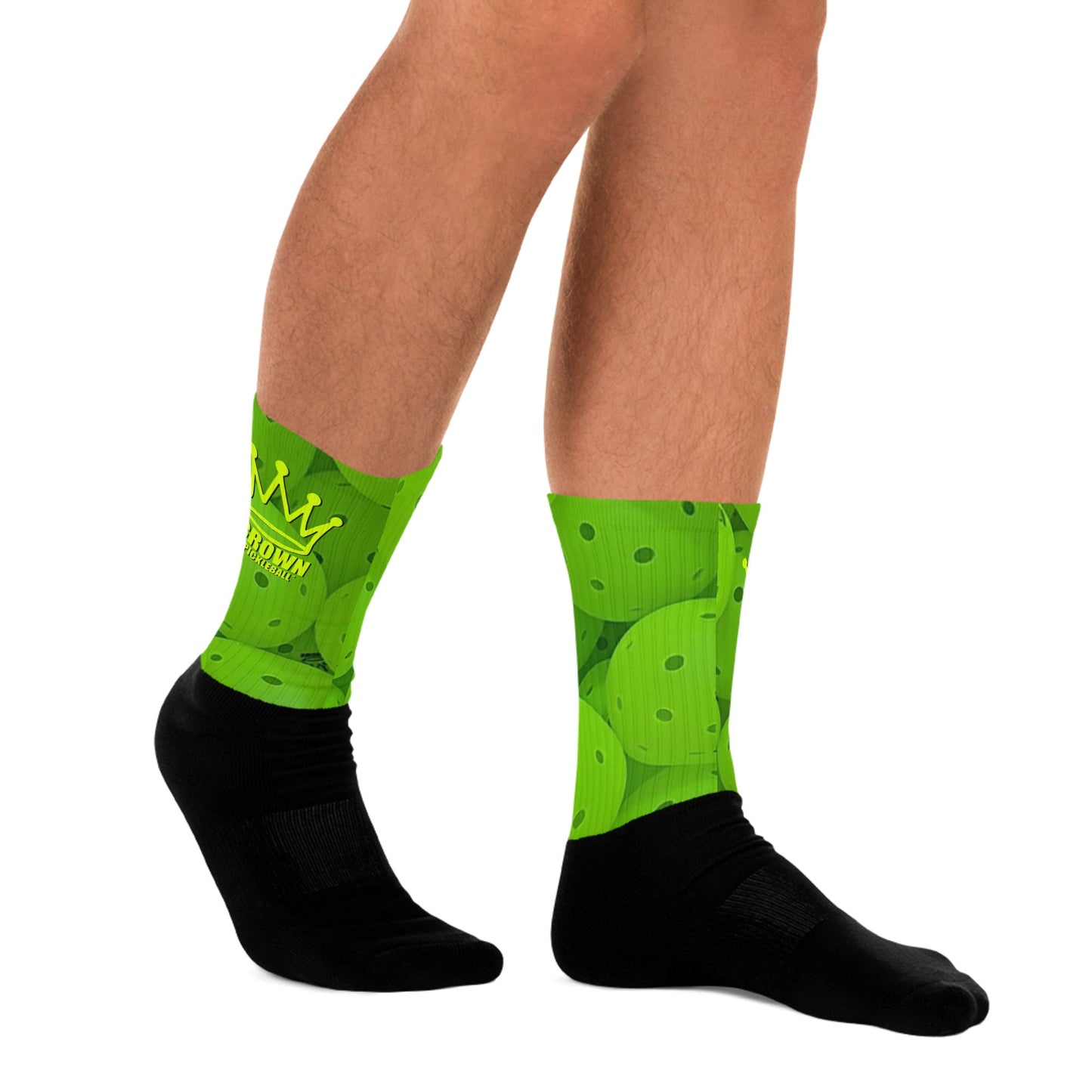 A person wears Crown Pickleball’s CROWN PICKLEBALL Performance Athletic Crew Socks featuring a green upper with crown logo and pickleball paddle and ball design, plus solid black foot and ankle. Only lower legs and feet are shown.