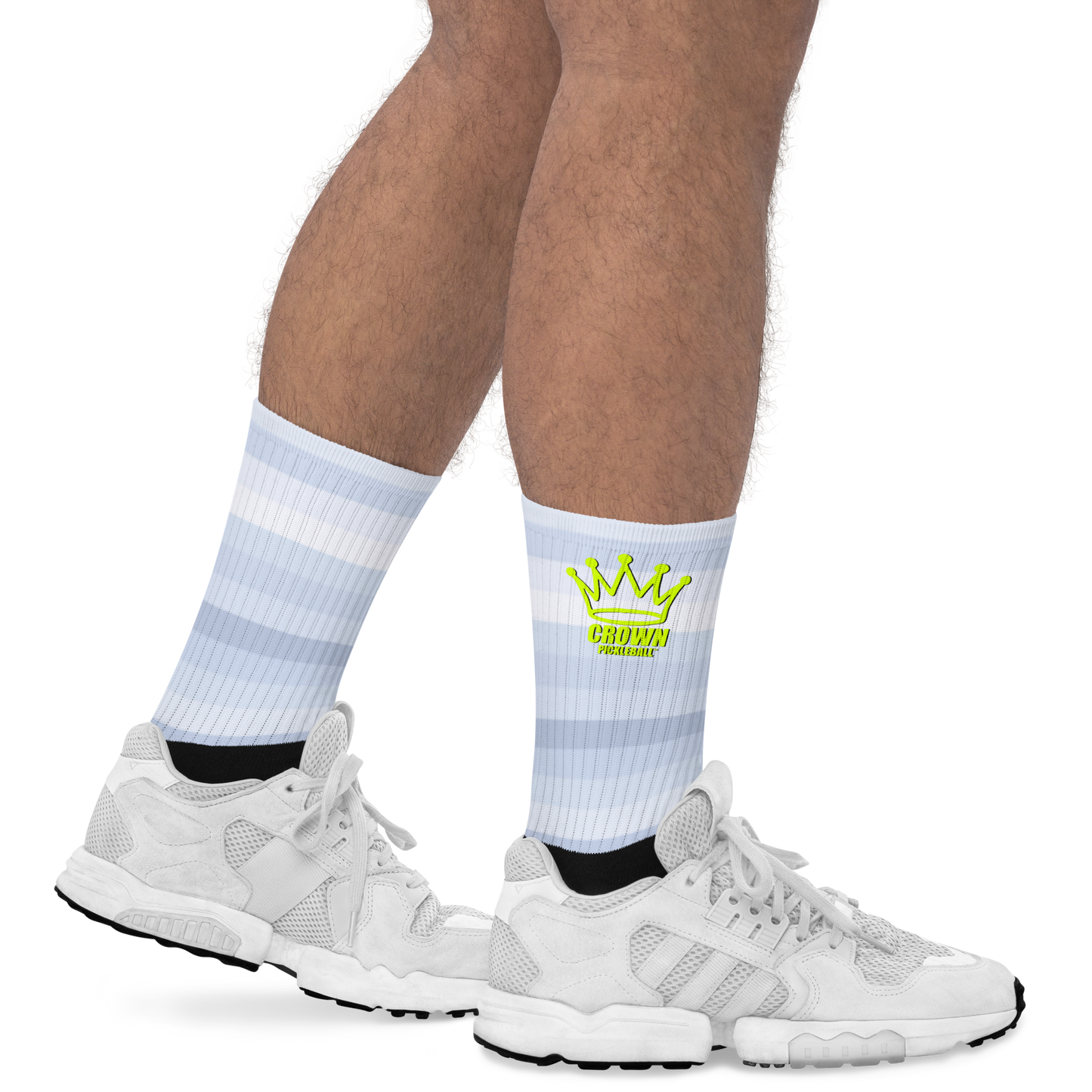 A person in white athletic shoes and Crown Pickleball Performance Athletic Crew Socks—with light blue stripes, a yellow crown graphic, and "CROWN COLLECTIVE" in green—stands legs apart on a white background; only lower legs and feet are visible.
