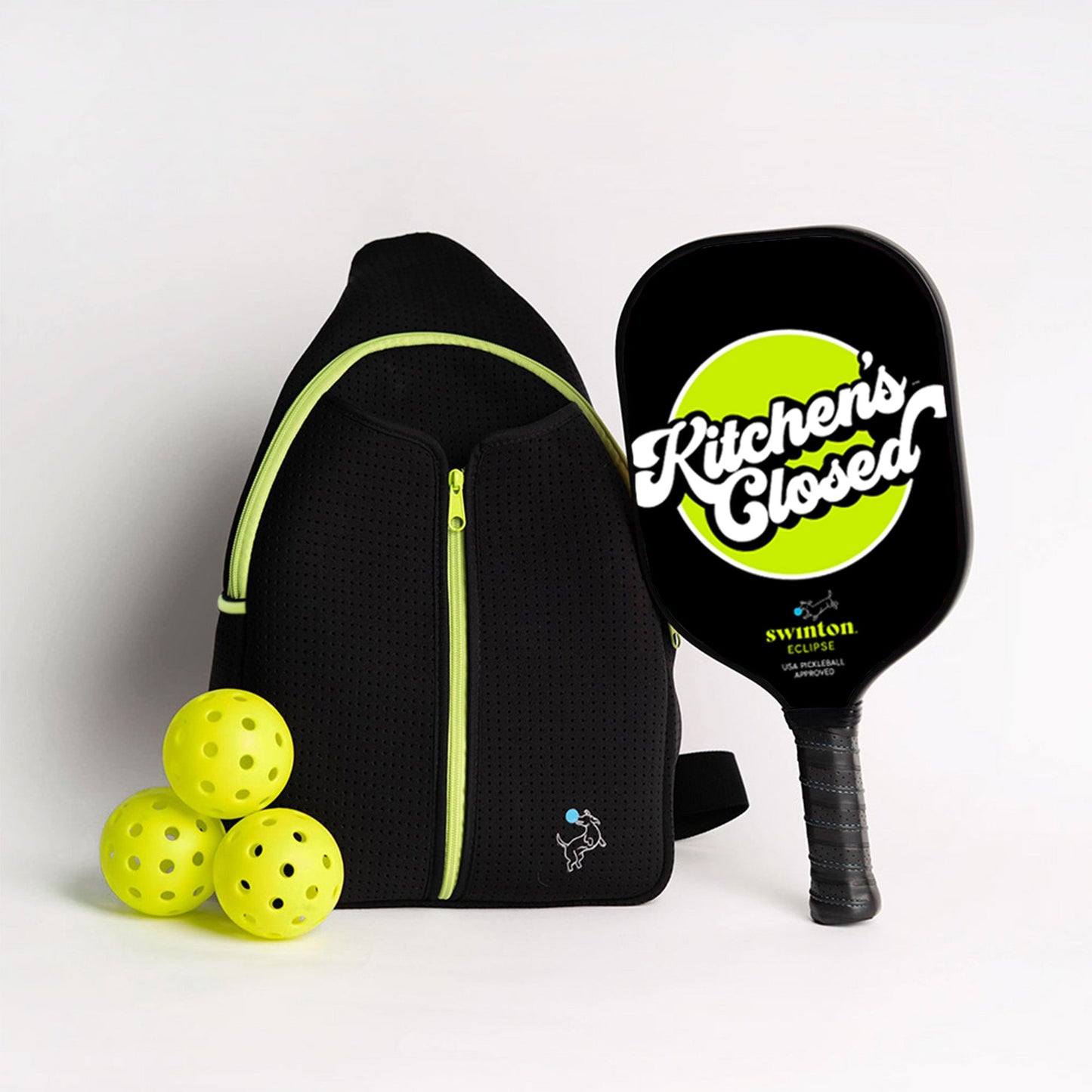The Swinton Pickleball Starter Bundle - Black Sling Bag Combo includes a black backpack with neon yellow zippers, a paddle reading “Kitchen’s Closed” on a neon yellow circle, and three yellow pickleballs—perfect for gifting or storing your gear.