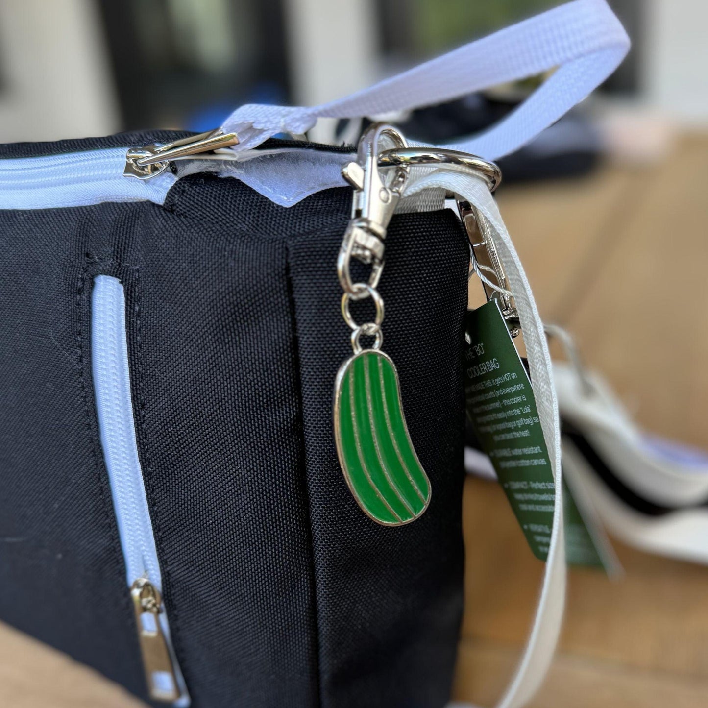 A close-up of the Fresh Pickle Insulated Cooler Bag in black with a light blue zipper and a green pickle keychain. Sunglasses rest on wood in the blurred background, suggesting court essentials.