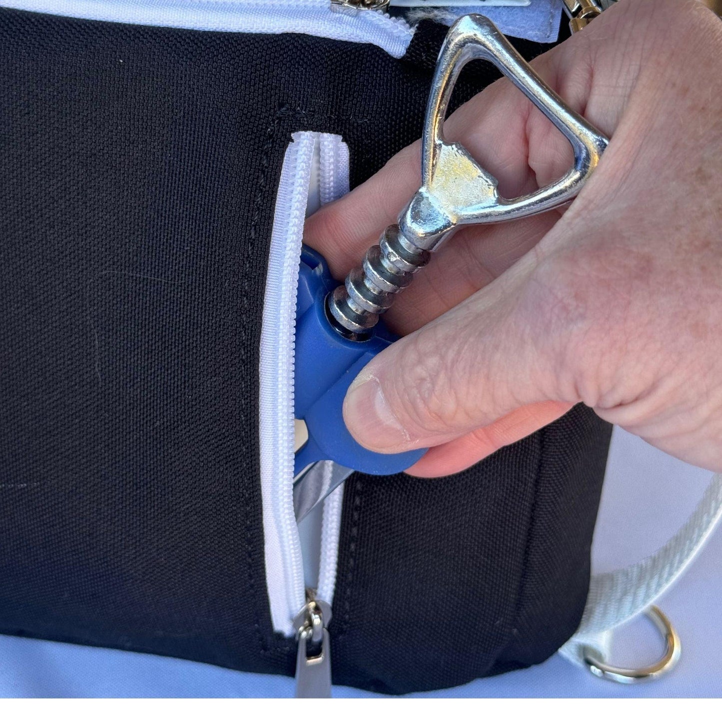 A hand pulls a metal corkscrew with a blue handle from the zippered pocket of the Fresh Pickle Insulated Cooler Bag, featuring a silver strap and resting on a white surface—ideal for carrying court essentials in style.