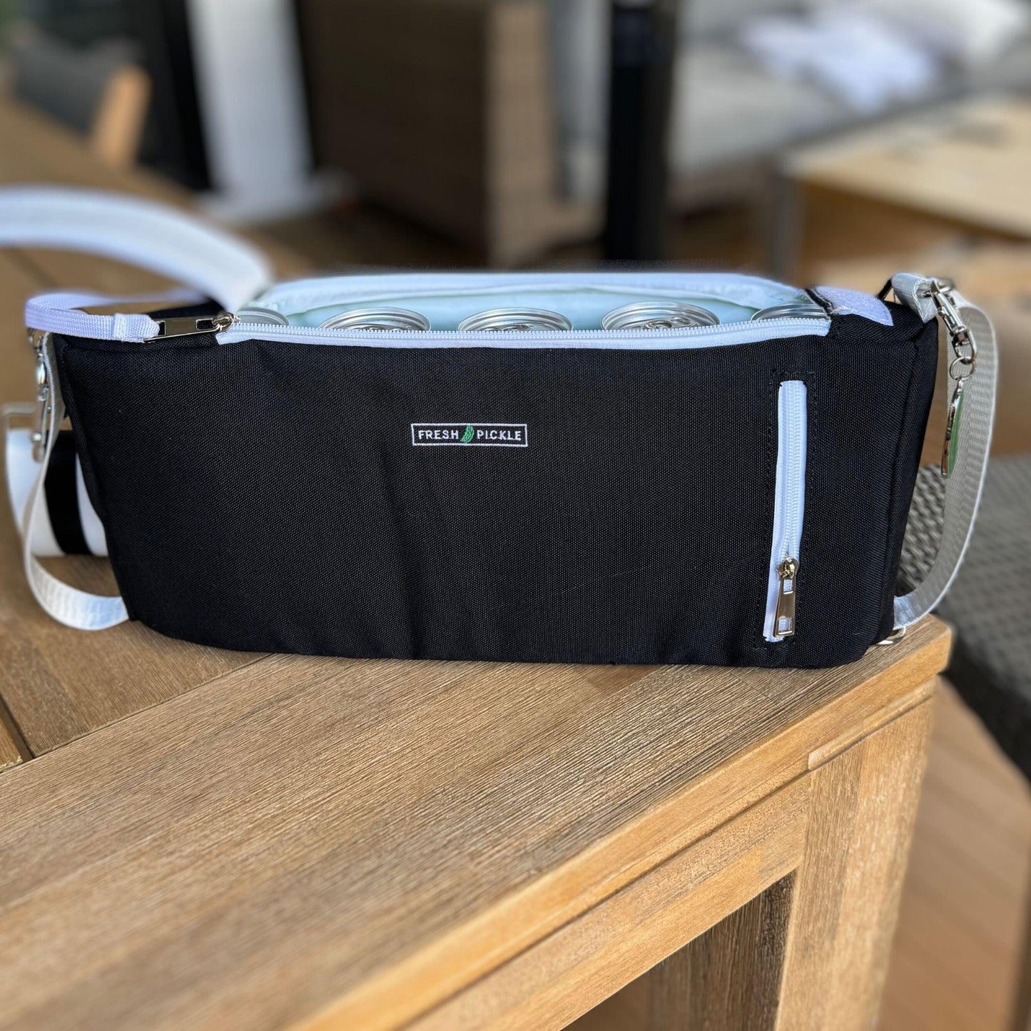 A black Fresh Pickle Insulated Cooler Bag with a light blue zipper sits on a wooden table. Four glass baby bottles are inside the soft-sided cooler, which has a white adjustable shoulder strap and small front zipper pocket.