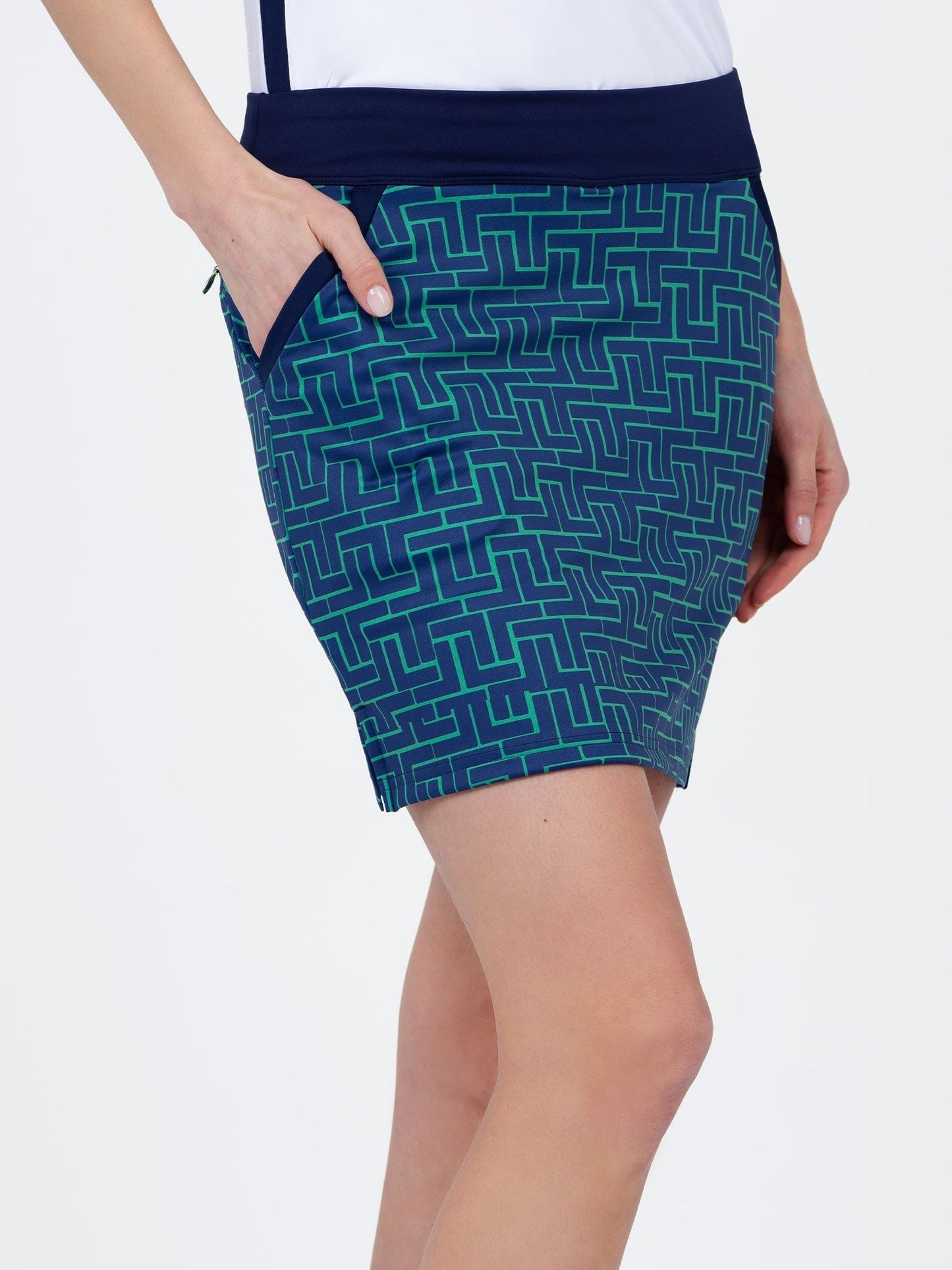 A person stands with one hand in the pocket of the inPhorm NYC Blake 17" Print Golf Skirt - Ink Tessel, a blue above-the-knee skirt with a green geometric pattern, made from recycled polyester. They pair it with a tucked-in white top against a plain white background.