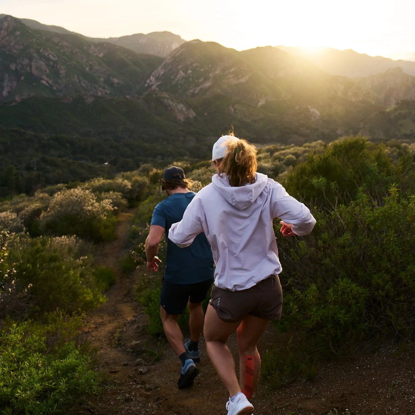Two people trail run at sunset through green hills, wearing athletic clothes and caps. One has a white hoodie, both ready for adventure and comfort with KT Health Blister Treatment Patch by KT Tape during their serene, active journey.