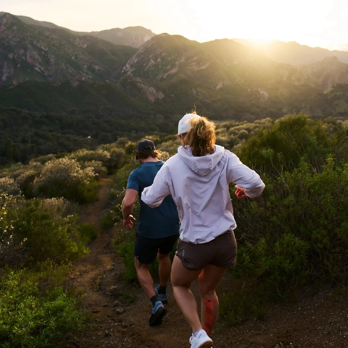 Two people trail run down a dirt path through green hills, sunlight streaming over mountains. Both look energized and ready for adventure with KT Tape KT Health Blister Treatment Patches to keep their run comfortable and blister-free.