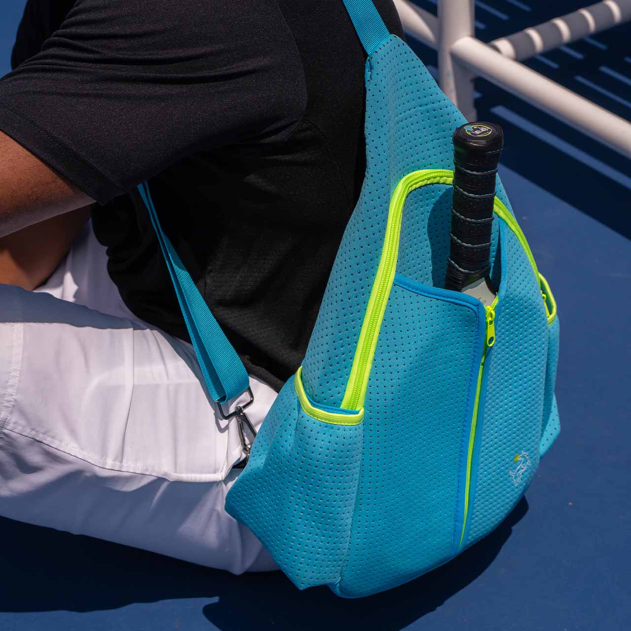A person in a black shirt and white shorts sits on a blue tennis court, carrying the Swinton Pickleball Sling Bag - Blue with a yellow zipper and racquet in the outer pocket. A white bench is partially visible nearby.