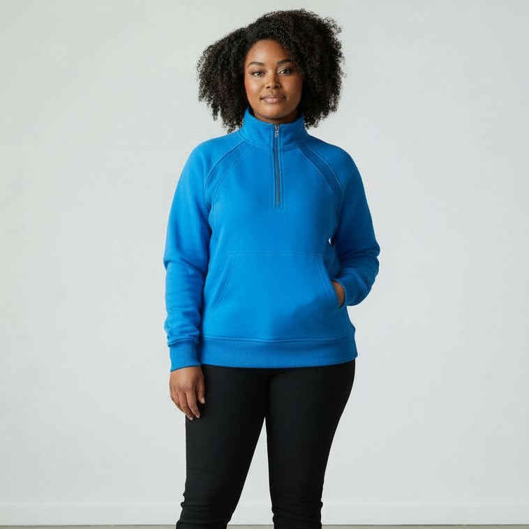 A woman with curly hair faces forward, wearing a bright blue DYM Athletics Cozy Up Full Length 1/2 Zip Sweatshirt and black pants. She has one hand in her pocket, a relaxed expression, and stands against a plain, light-colored background.