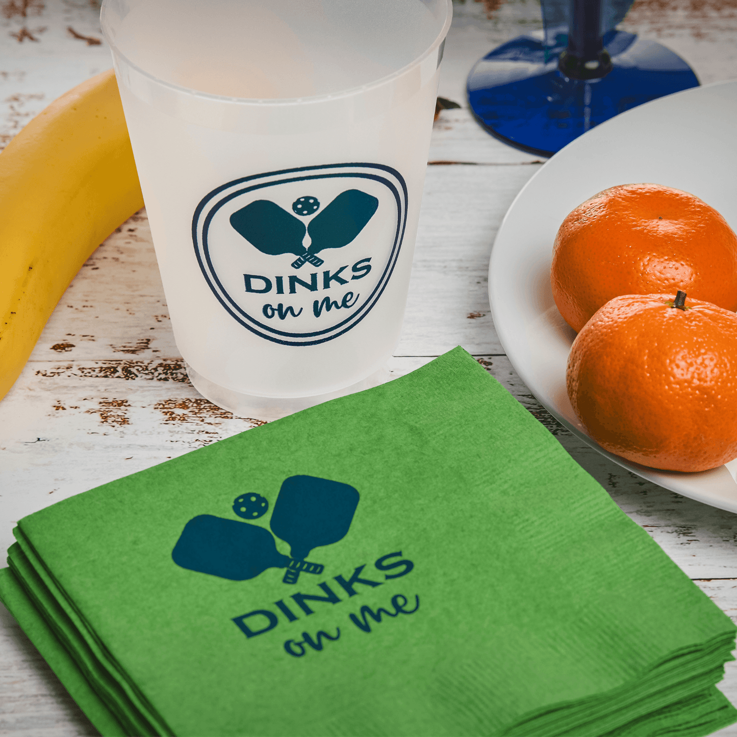 A Fresh Pickle Pickleball Roadie Cup and green napkins, both with blue pickleball paddles and “DINKS on me,” sit near a yellow banana, two oranges on a white plate, and a blue-stemmed glass atop a white wooden surface.