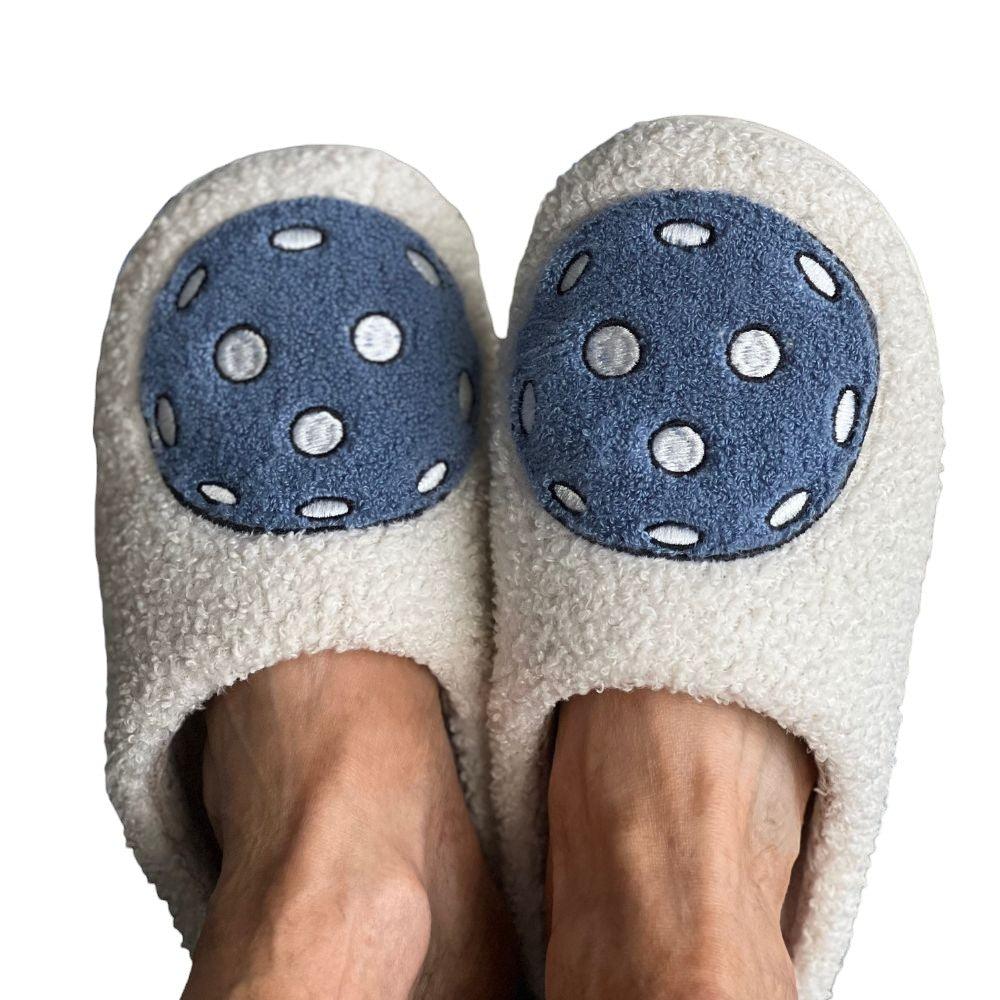 A pair of feet wear Fresh Pickle's Pickleball Slippers—fuzzy, cream-colored comfort with large blue-and-white polka dot patches. The plain white background keeps focus on these cozy, standout slippers.