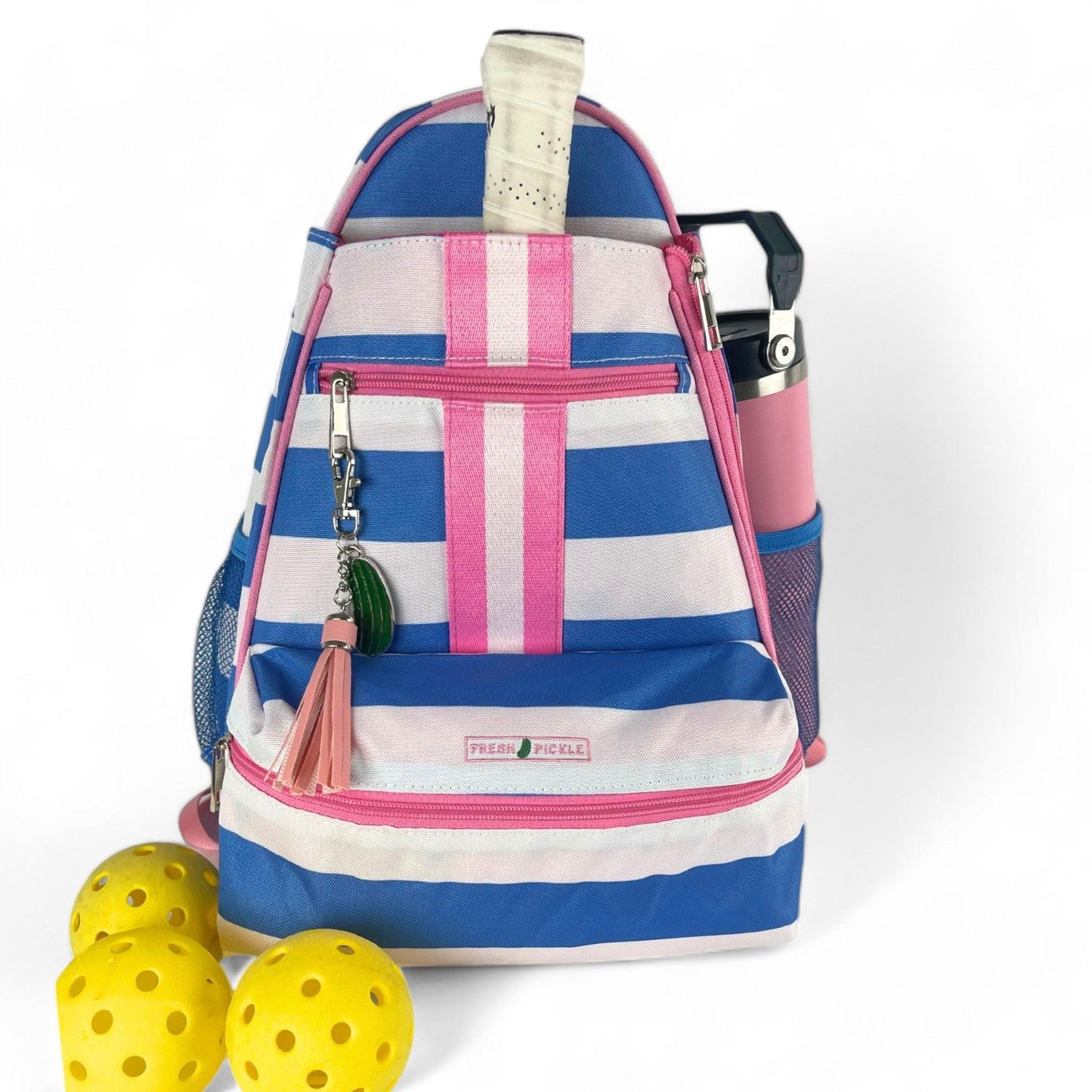 The Fresh Pickle Teddi Pickleball Backpack features blue, pink, and white stripes, a pink tassel keychain, and a water bottle in the side pocket. Three yellow perforated balls are placed in front on a white background.