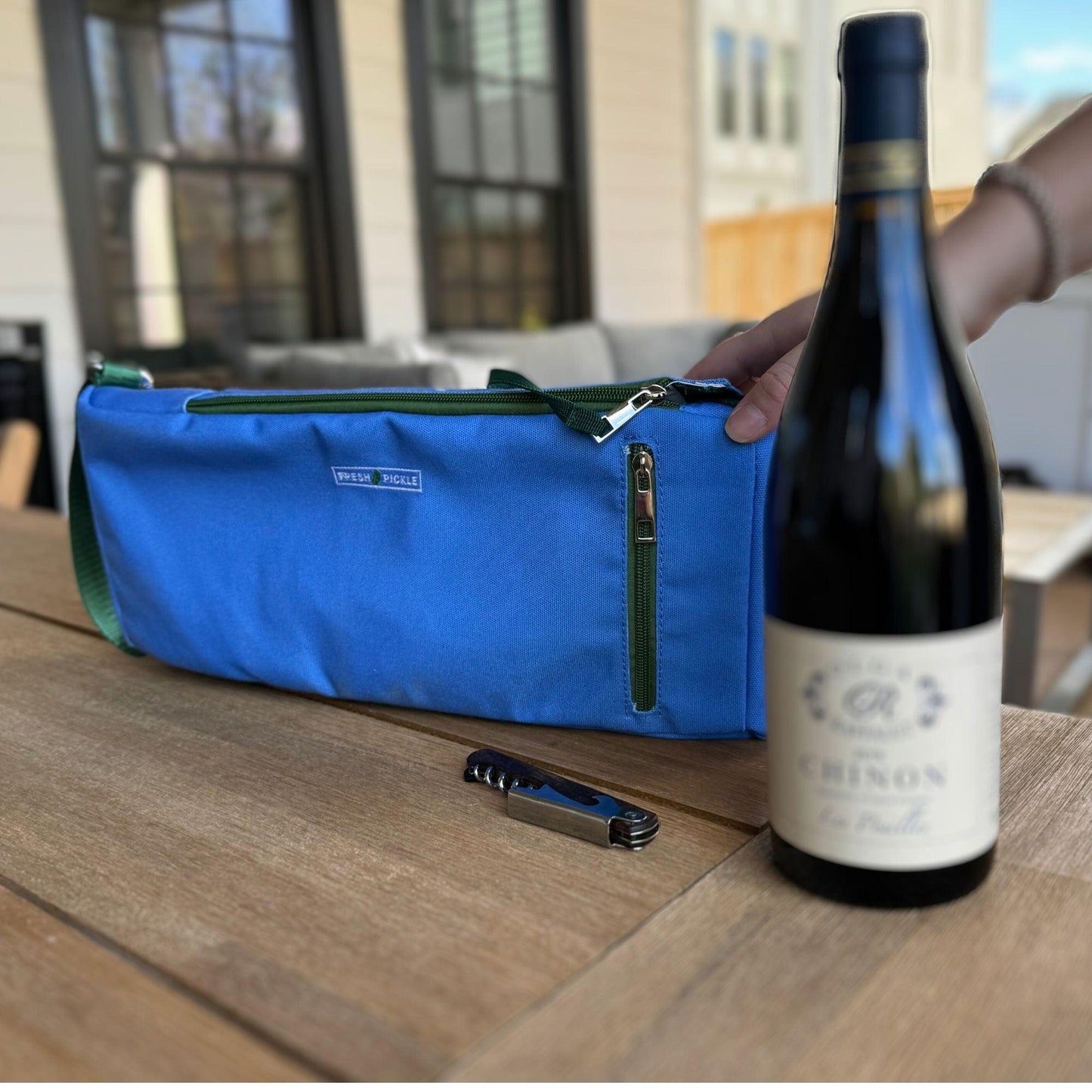 A hand holds a Fresh Pickle Insulated Cooler Bag with zippers on a wooden outdoor table. Next to it is a dark bottle of Chinon red wine and a closed corkscrew, with windows and a fence blurred in the background.